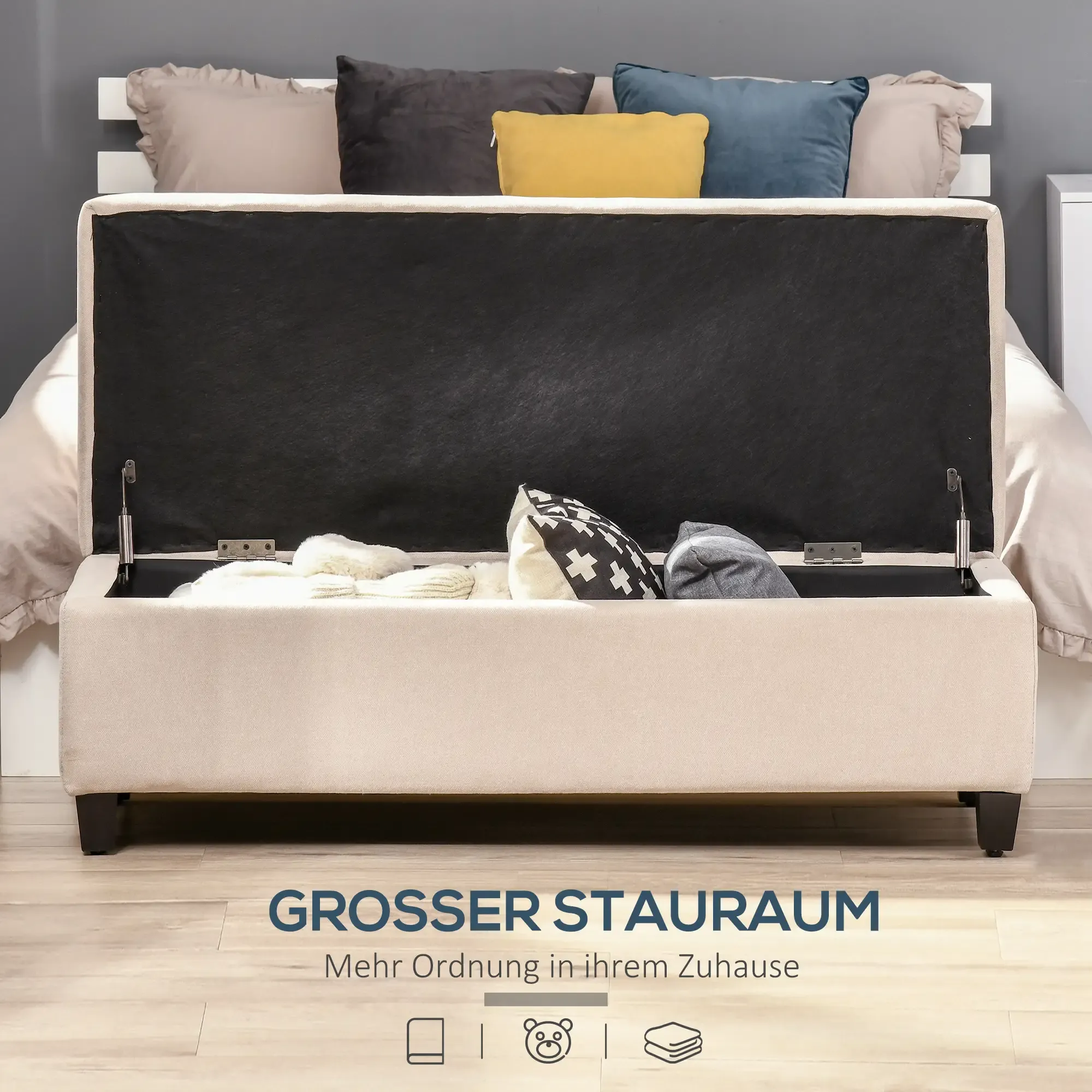 Beige Stauraum-Ottomane mit offenem Deckel. Kissen in Schwarz, Gelb und Blau liegen auf dem Bett. Weißes Kopfteil und Kissen sind sichtbar.
