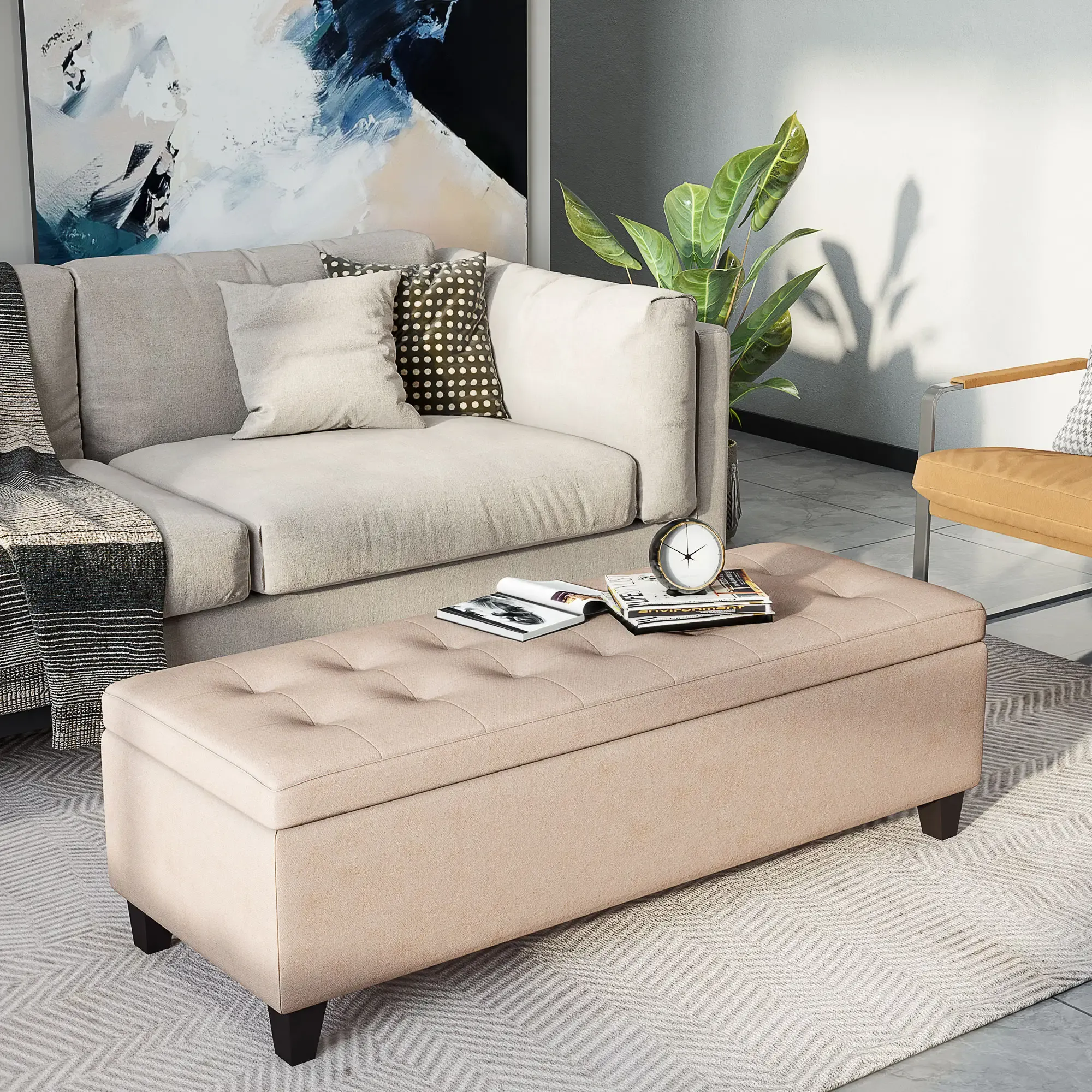 Eine beige Ottomane mit Büchern und einer Uhr steht vor einem grauen Sofa, Kissen und Kunstwerken. Rechts sind eine Pflanze und ein Stuhl.