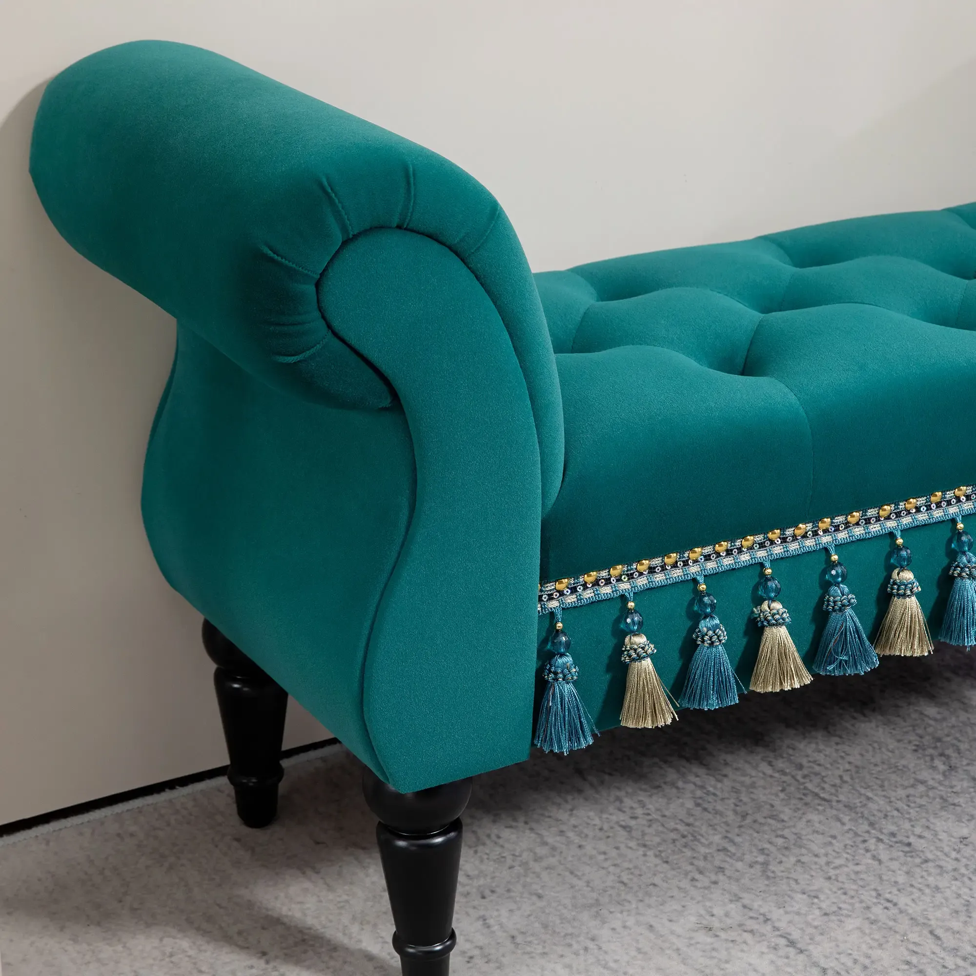 Eine teal farbene Samtbank mit abgerundetem Arm und getufteter Sitzfläche. Sie hat schwarze Beine und Quasten. Im Hintergrund eine Wand und Teppich.