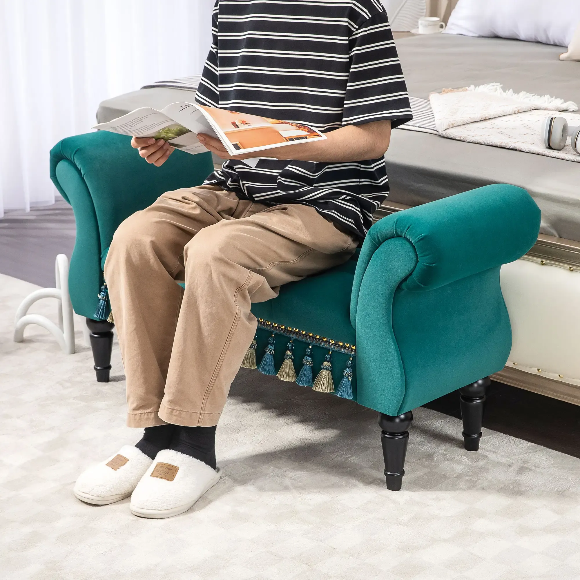 Eine Person liest eine Zeitschrift, während sie auf einer tealfarbenen Samtbank sitzt. Die Person trägt ein gestreiftes Hemd, beige Hose und Hausschuhe.