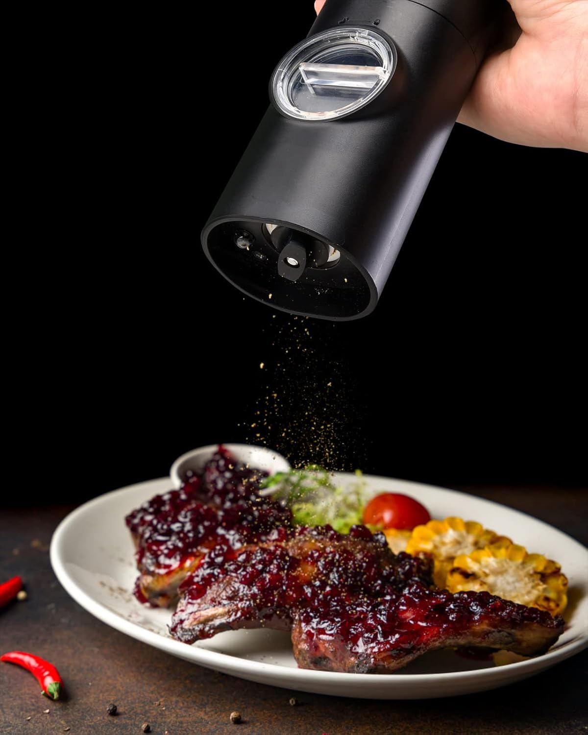 Eine Hand streut Gewürz auf BBQ-Rippchen, Mais und eine Tomate auf einem weißen Teller. Schwarzer Hintergrund.