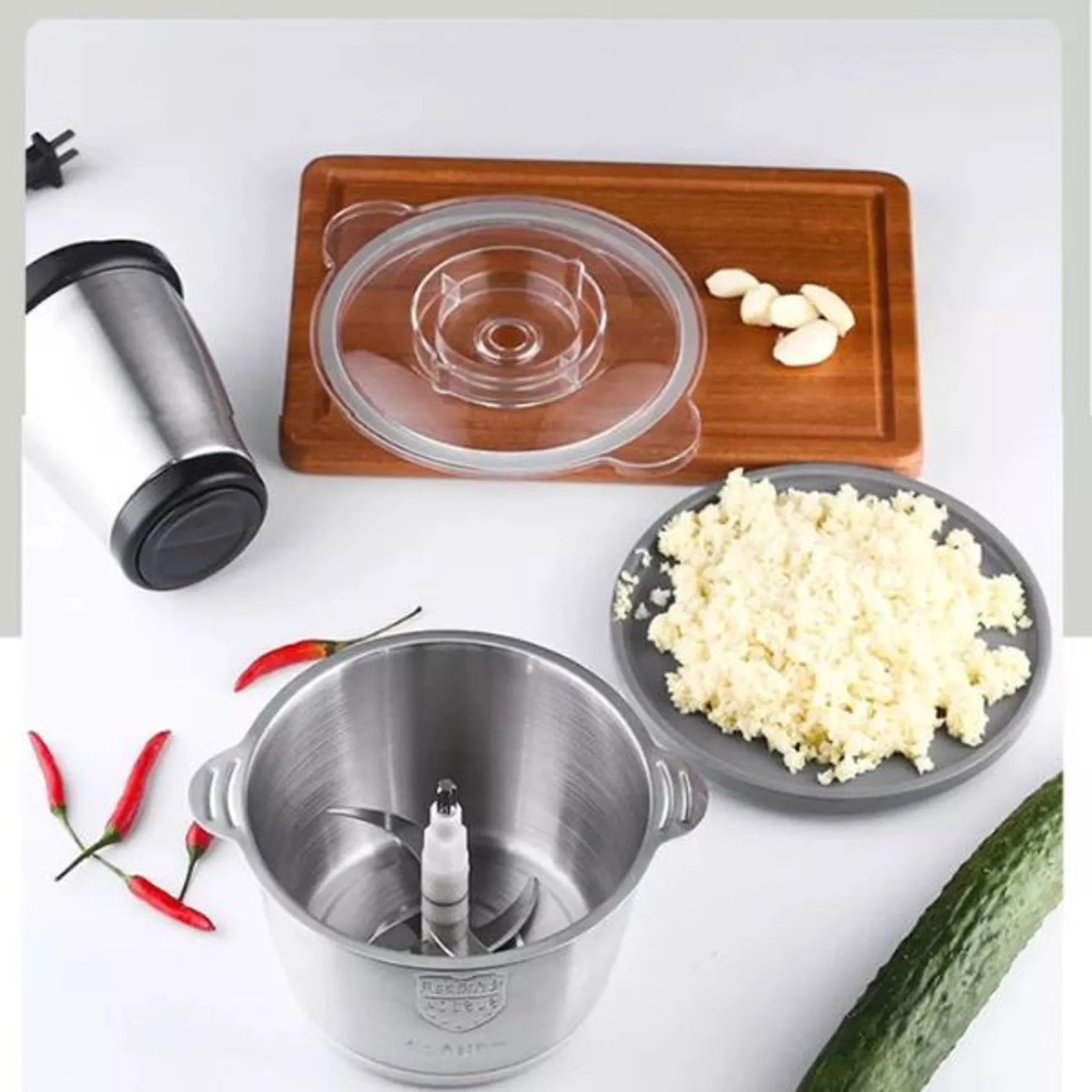 Ein Edelstahl-Lebensmittelprozessor mit Zutaten wie Knoblauch, Chilis und Gurke auf einer weißen Oberfläche.