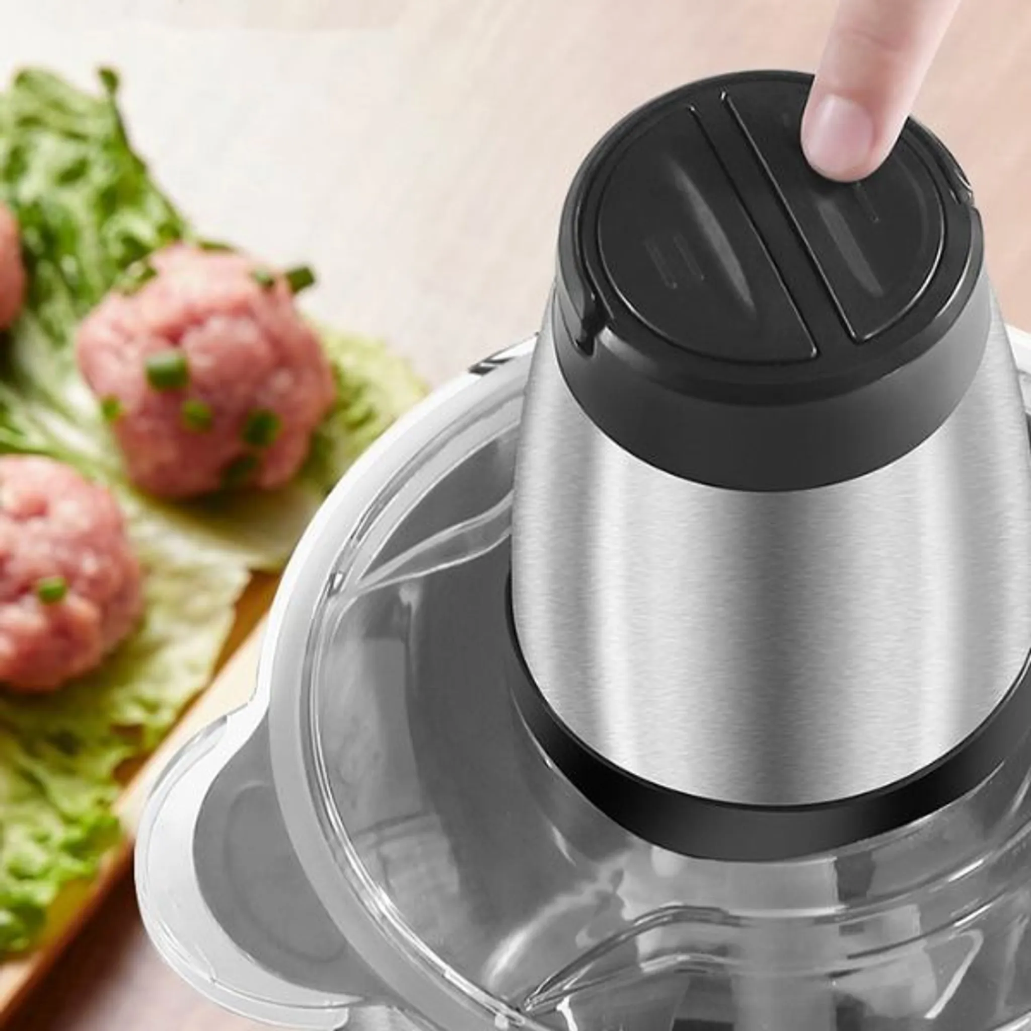 Eine Person drückt einen Knopf an einem Zerkleinerer. Rohe Fleischbällchen liegen im Hintergrund auf Salat.