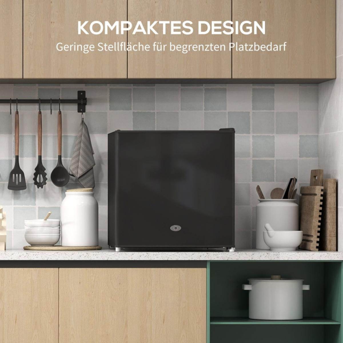 Ein schwarzer Minikühlschrank steht in einer Küche auf einer Arbeitsplatte. Utensilien und Gläser sind sichtbar. Die Schränke sind helles Holz und grün.