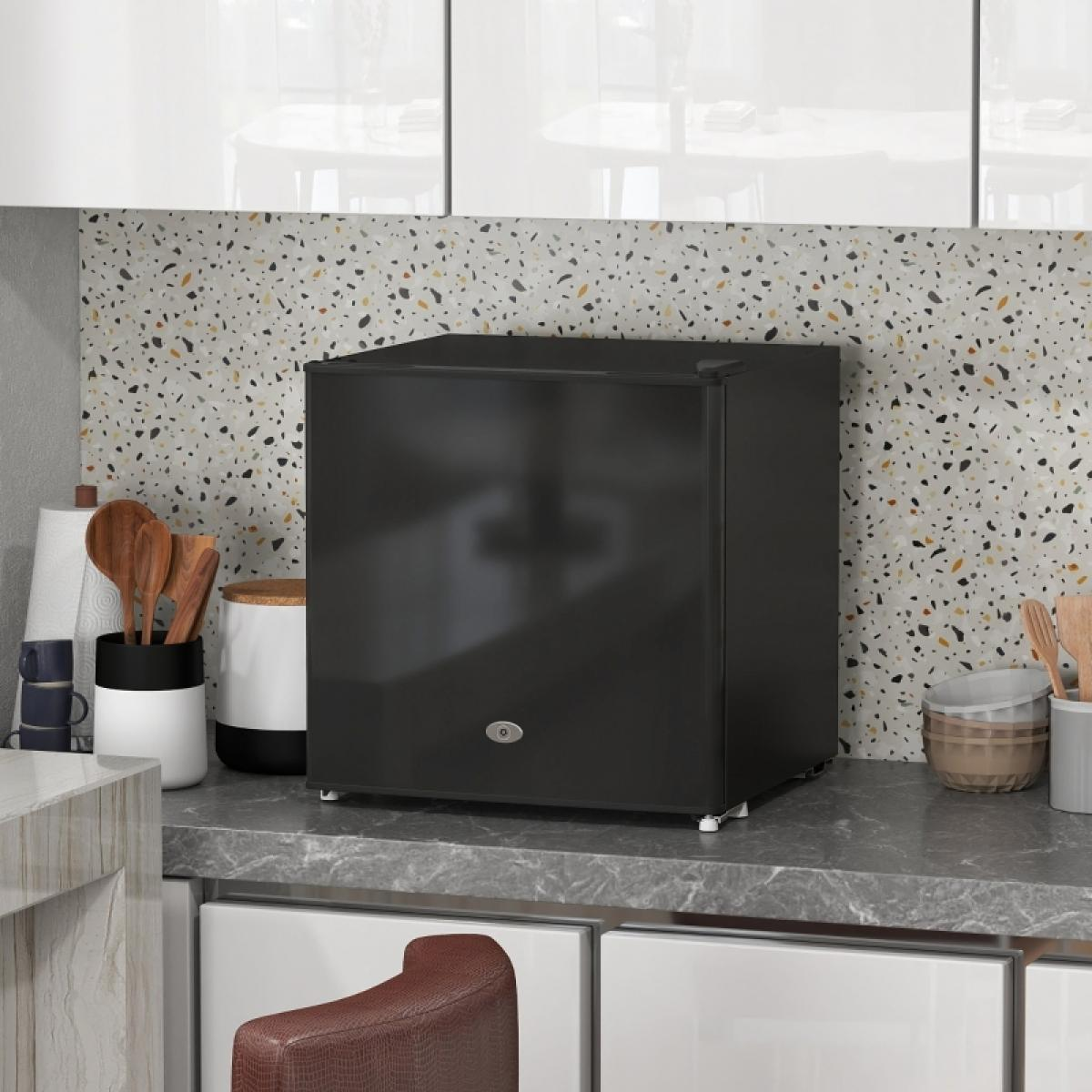 Ein schwarzer Mini-Kühlschrank steht auf einer Arbeitsplatte. Küchenutensilien und ein gemusterter Fliesenspiegel sind im Hintergrund. Ein brauner Sitz ist darunter.