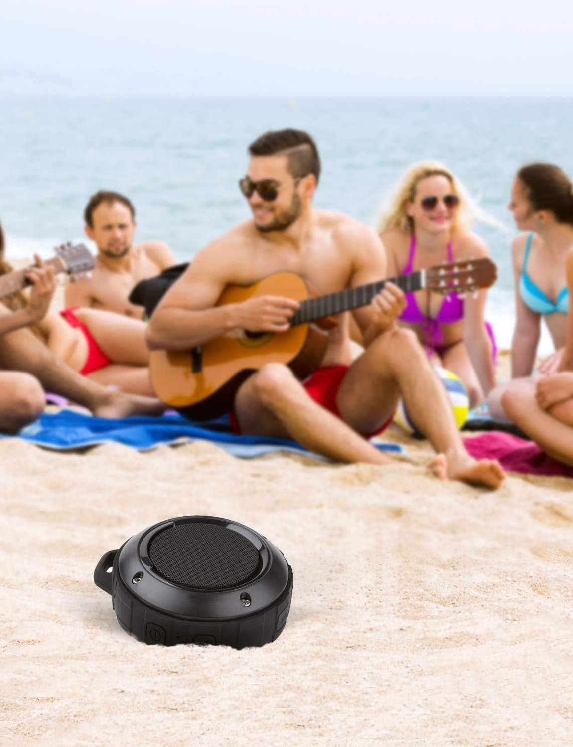 Ein schwarzer Lautsprecher steht auf Sand. Leute spielen am Strand Gitarre und genießen die Sonne. Das Meer ist im Hintergrund.