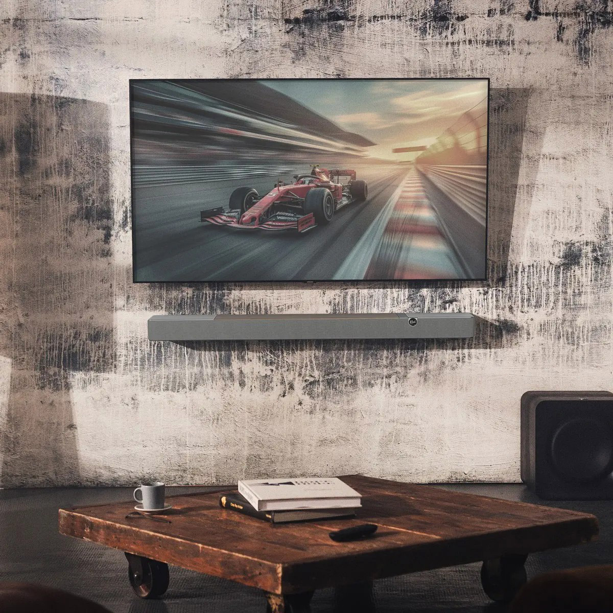 Ein Wohnzimmer mit einem Fernsehgerät, das ein Rennauto zeigt. Soundbar darunter. Couchtisch mit Büchern und einer Tasse. Ein Subwoofer rechts.