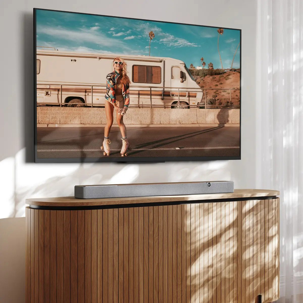 Ein Fernseher zeigt eine Person beim Rollschuhlaufen. Eine Soundbar liegt auf einem Holzschrank. Sonne scheint durch einen weißen Vorhang.