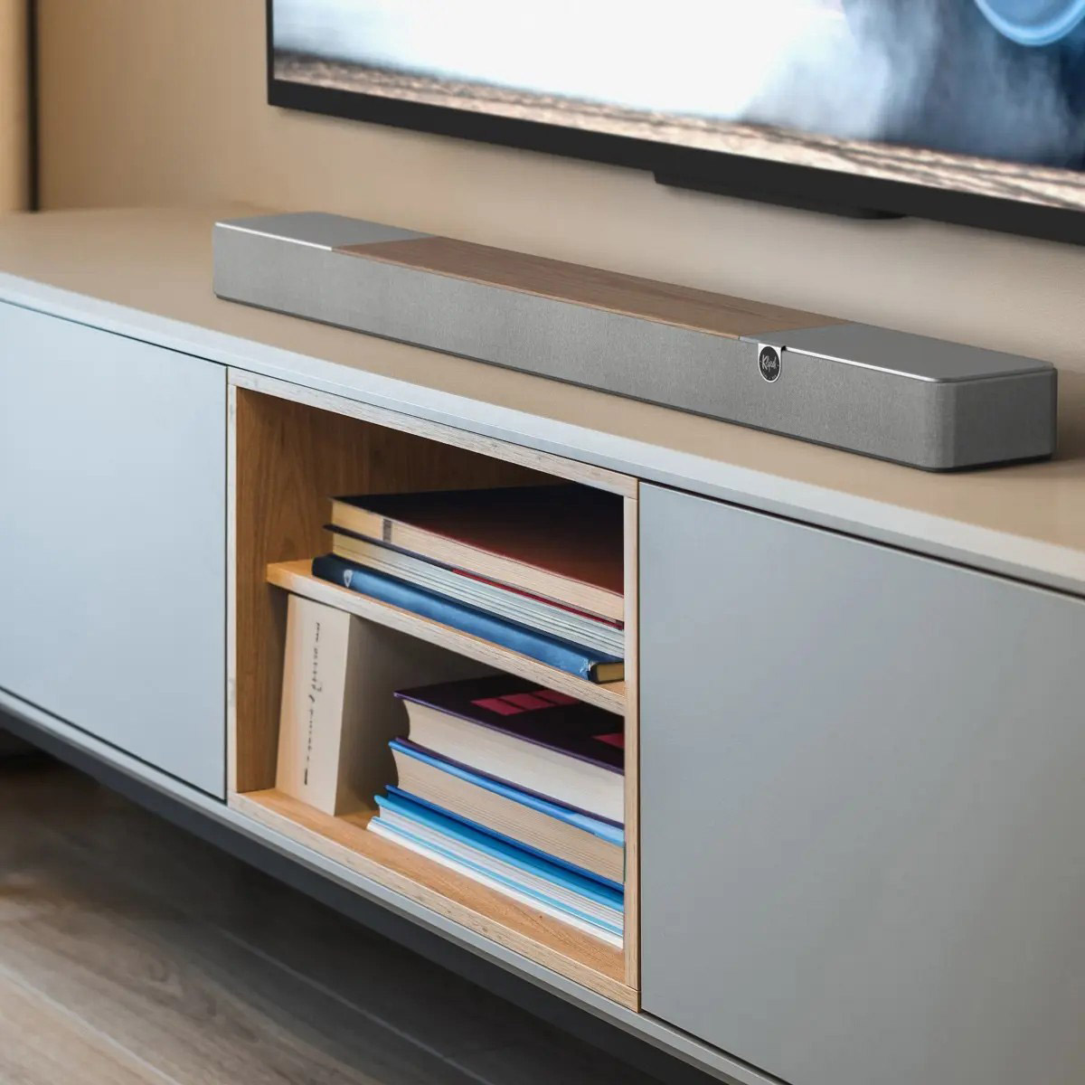 Eine Soundbar liegt auf einem Schrank mit Büchern im offenen Fach. Darüber befindet sich ein Fernseher. Die Soundbar hat silberne Enden und einen Holzabschnitt.