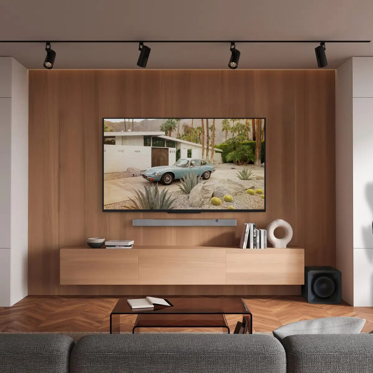 Wohnzimmer mit TV mit Auto. Ein Holzschrank enthält Bücher und Deko. Schienenbeleuchtung beleuchtet die Szene. Vorne ein graues Sofa.