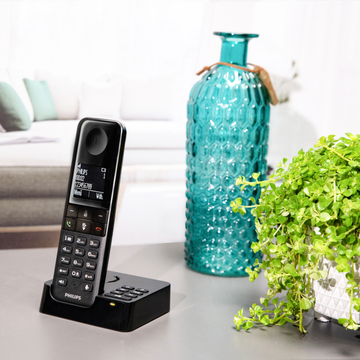 Un téléphone sans fil sur un chargeur. Un vase en verre turquoise et une plante verte en arrière-plan. Le téléphone affiche des chiffres.