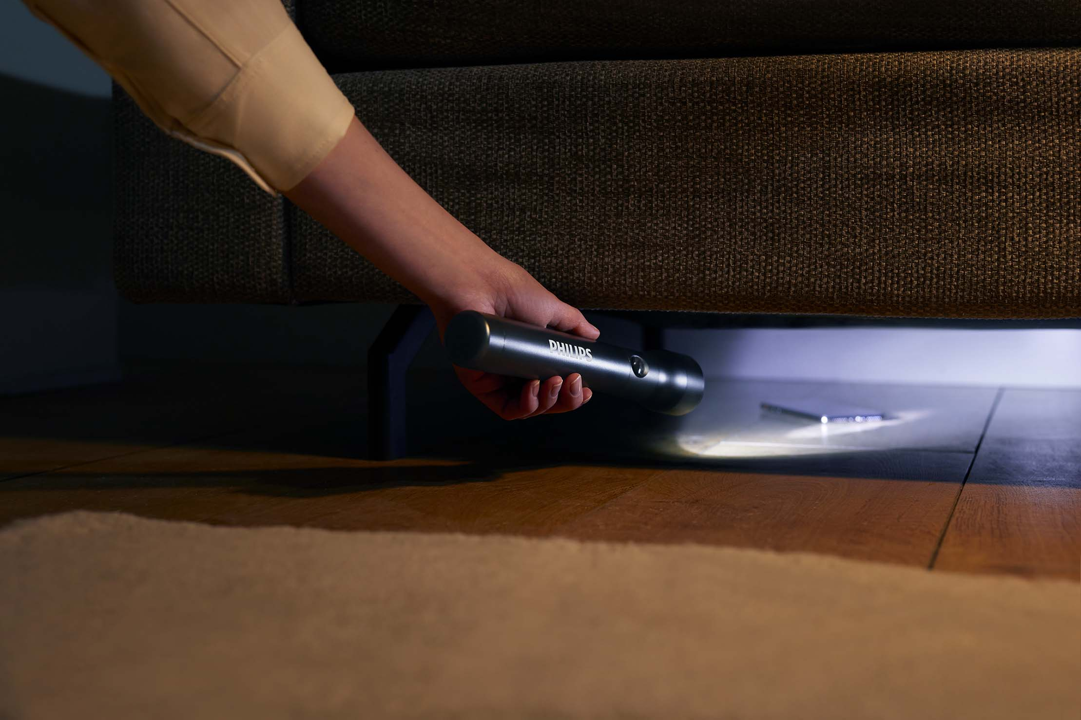 Una mano tiene una torcia Philips sotto un divano, illuminando lo spazio. Un telefono è anche sotto il divano, su un pavimento di legno.