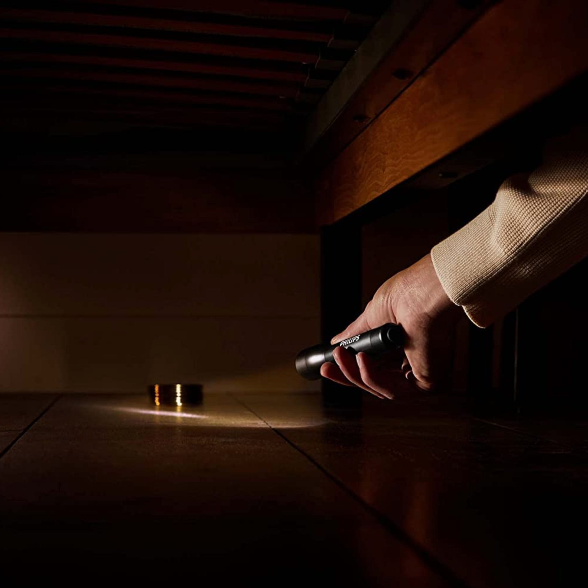 Una mano con una torcia Philips illumina sotto un letto. Un piccolo oggetto è illuminato sul pavimento.