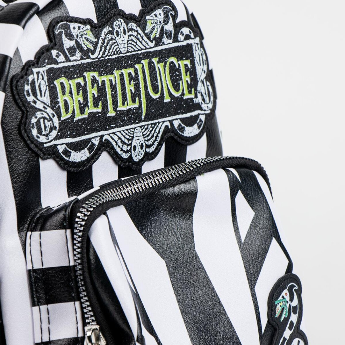 Mochila a rayas blancas y negras con un parche de 'Beetlejuice'. El parche tiene calaveras y texto verde. Cremalleras y un parche pequeño son visibles.