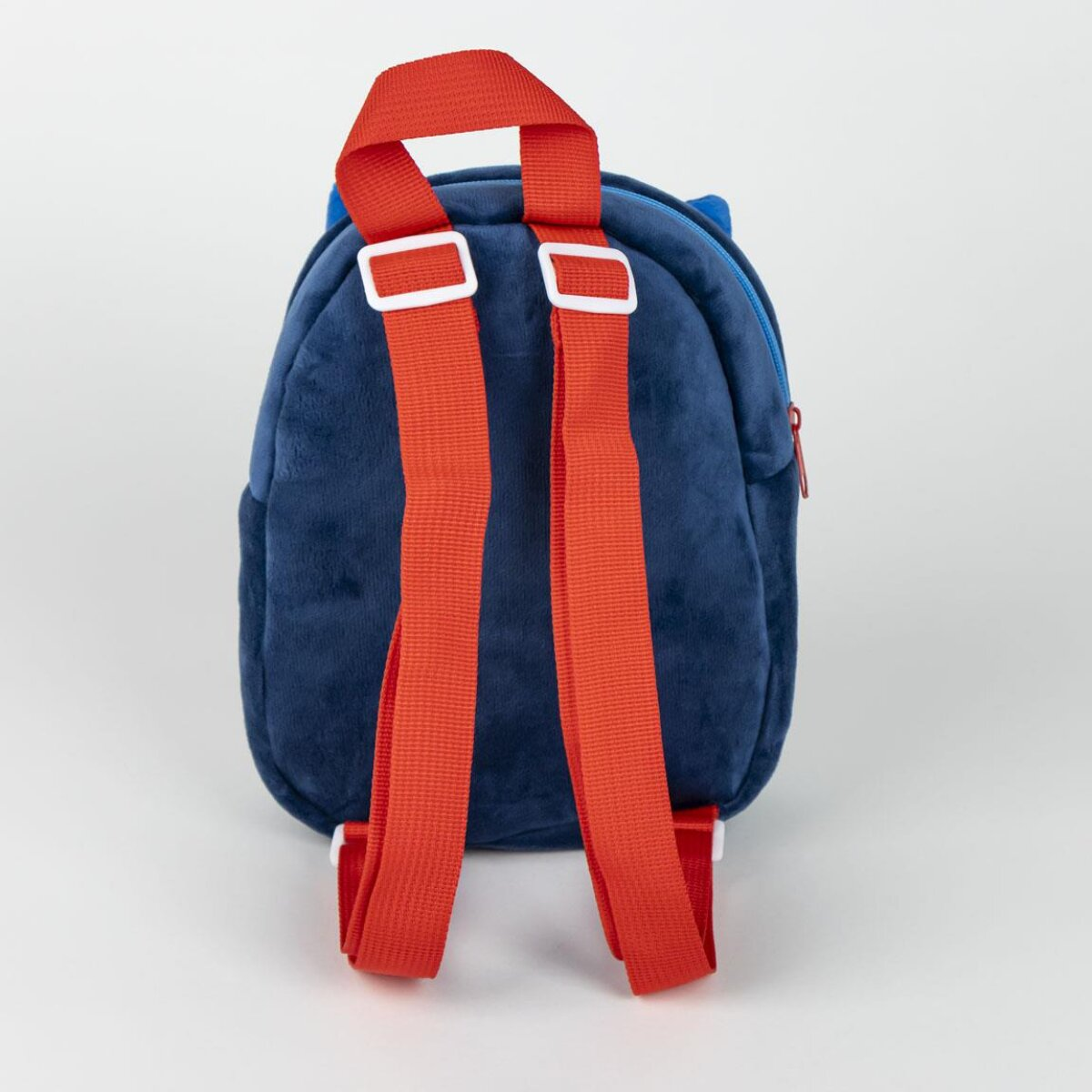 Una mochila de felpa azul marino con correas rojas brillantes y hebillas blancas. El fondo es blanco liso.