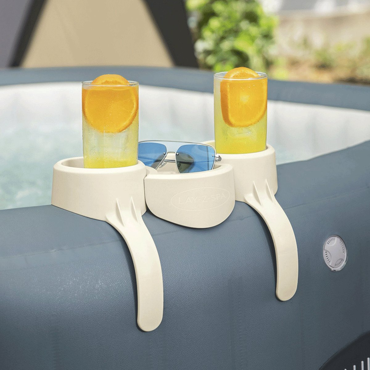 Twee drankjes met sinaasappelschijfjes, een zonnebril en een accessoire voor een bubbelbad. De setting is buiten met wazig groen op de achtergrond.