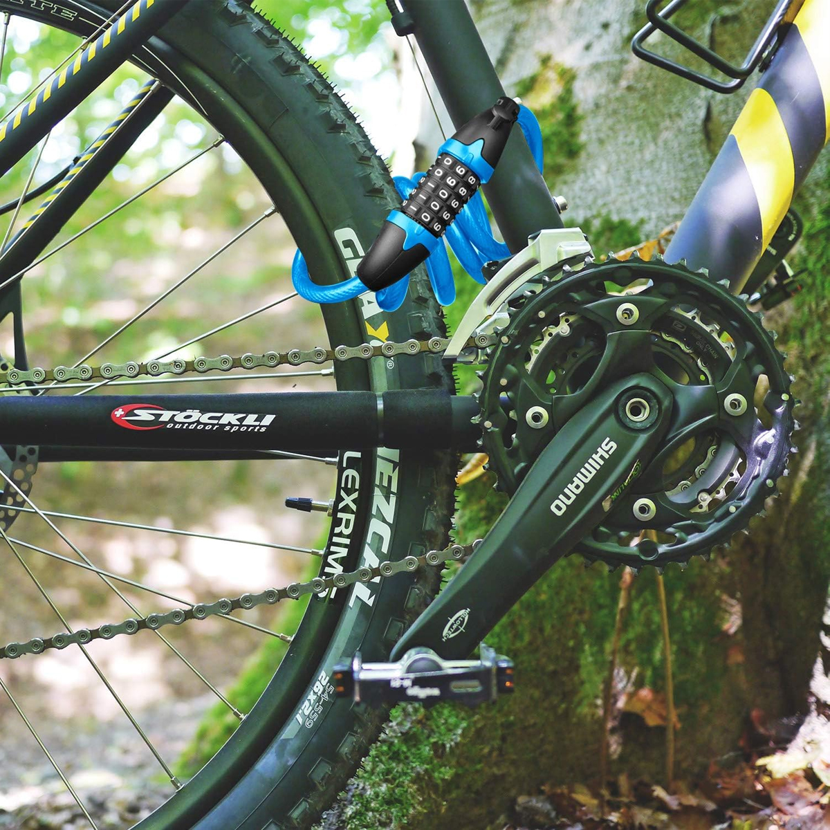 Ein Fahrradrad ist mit einem blauen Kabelschloss verschlossen. Das Fahrrad hat einen schwarzen Rahmen, Gänge und eine Shimano-Kurbelgarnitur. Der Hintergrund sind verschwommene Bäume.