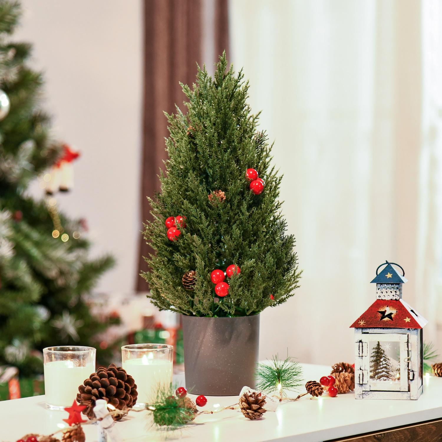 Ein kleiner Weihnachtsbaum in einem Topf, rote Beeren, Kerzen, Tannenzapfen und eine Laterne stehen auf einem weißen Tisch.