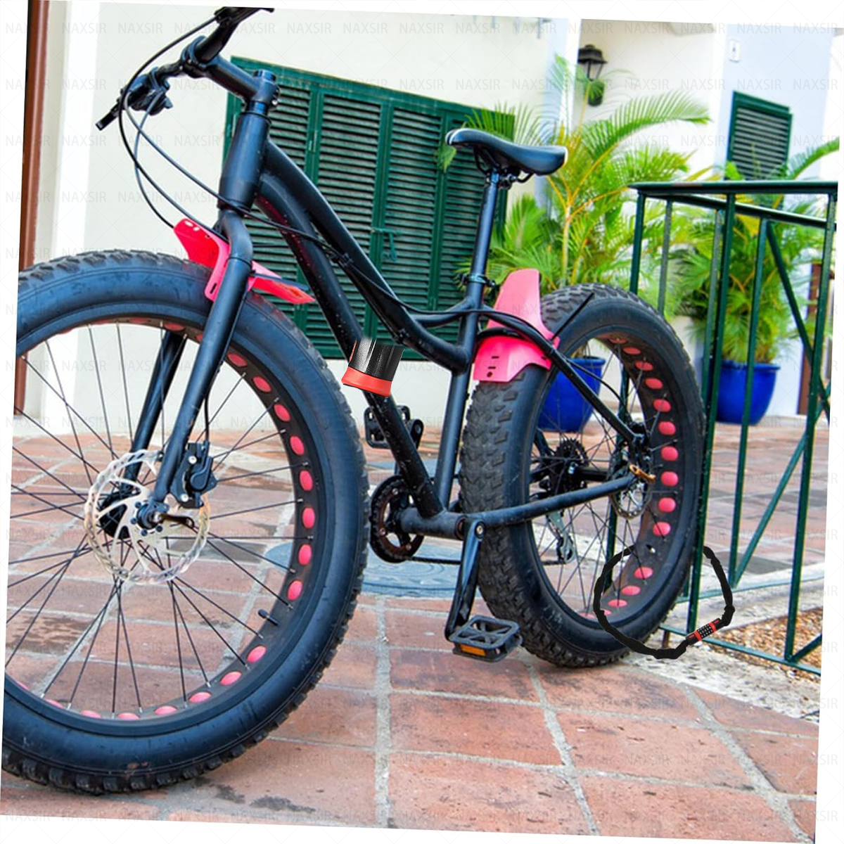 Ein schwarzes Fatbike mit roten Akzenten ist an einem Geländer befestigt. Der Hintergrund zeigt ein Gebäude mit grünen Fensterläden und Pflanzen.