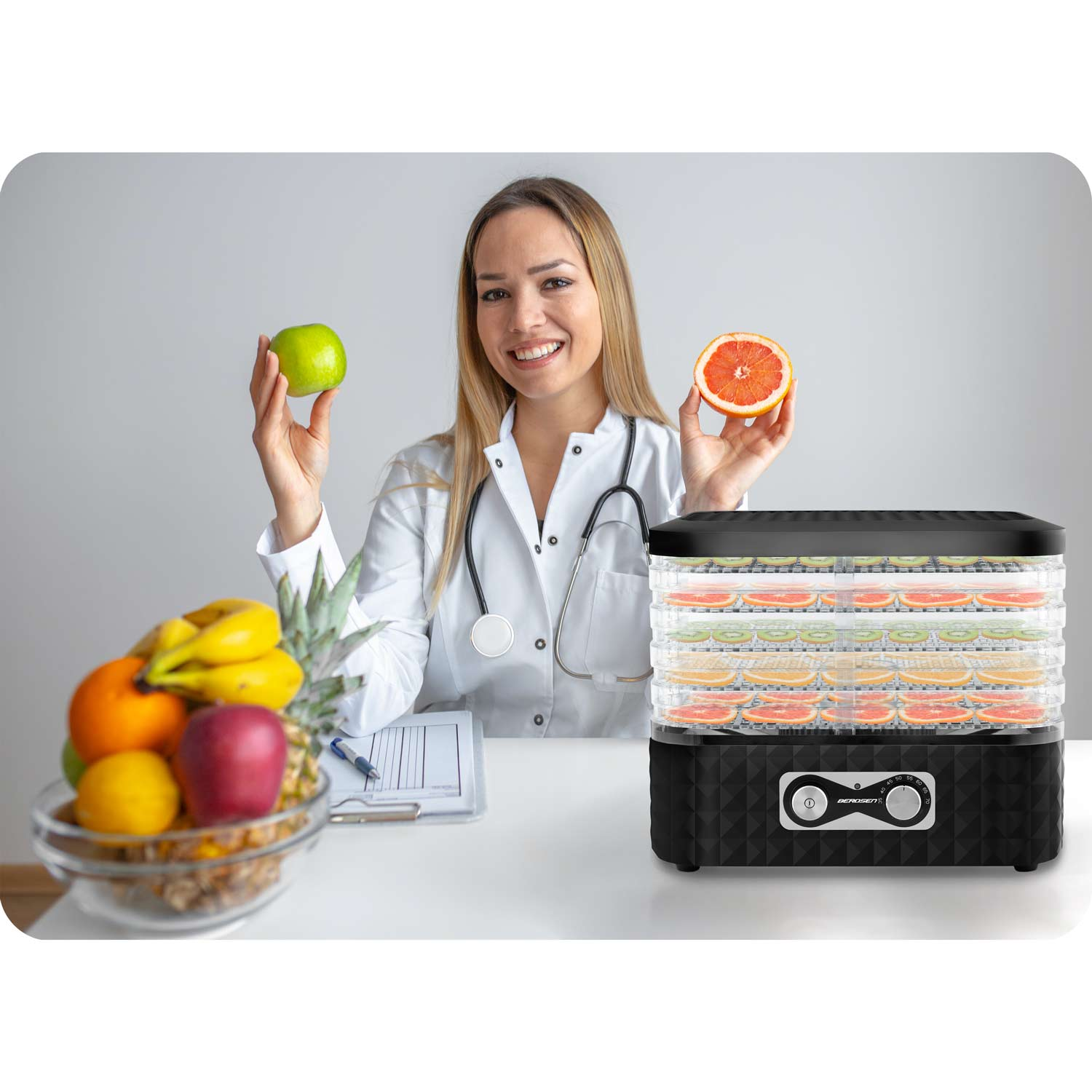 Uśmiechnięty lekarz trzyma zielone jabłko i grejpfruta. Suszarka do żywności i miska owoców stoją na białym stole.