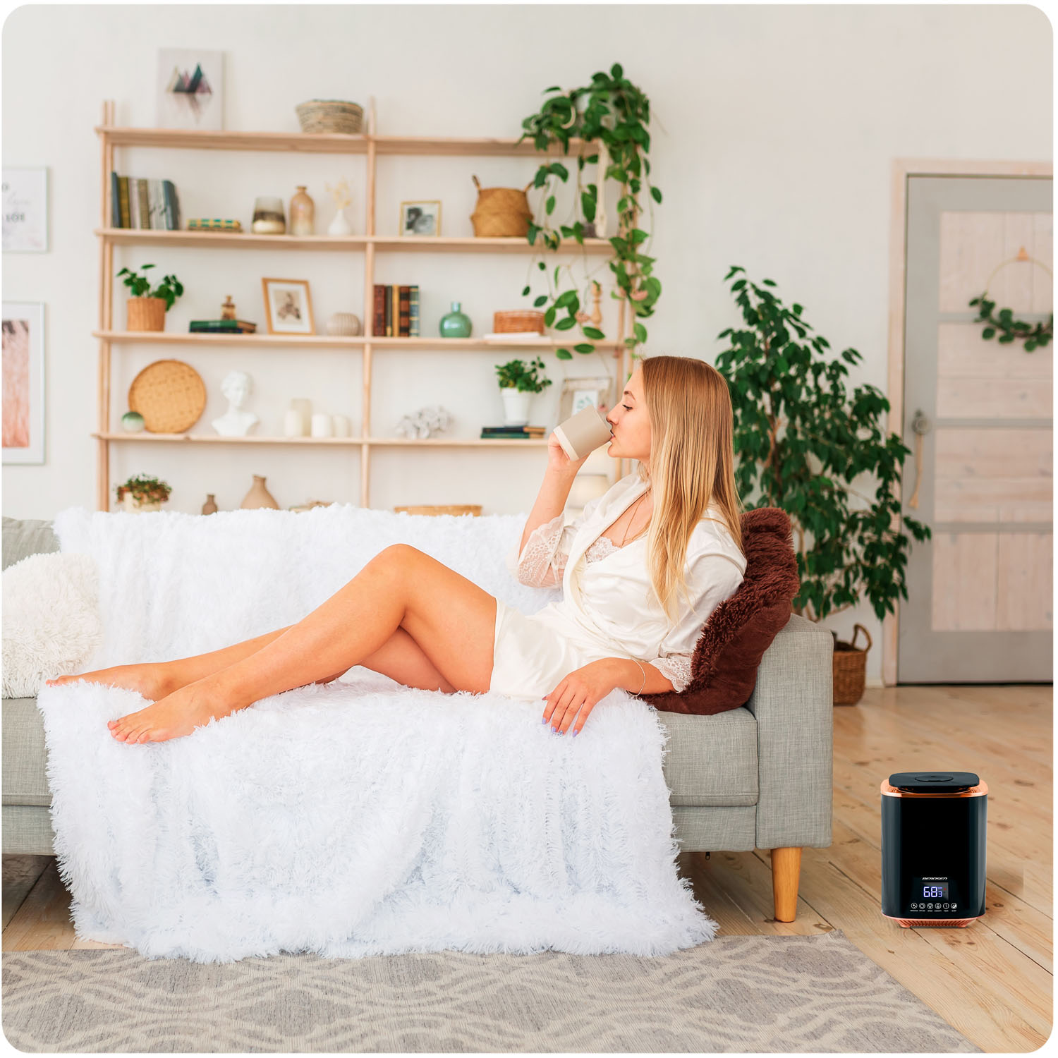 Kobieta pije z kubka, leżąc na kanapie. Nawilżacz powietrza stoi w pobliżu. Półki z dekoracjami znajdują się w tle.