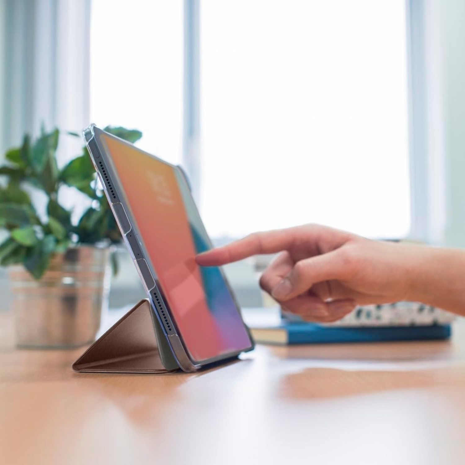 Eine Hand berührt einen Tablet-Bildschirm. Das Tablet steht auf einem Ständer. Eine Pflanze und Bücher sind im Hintergrund.