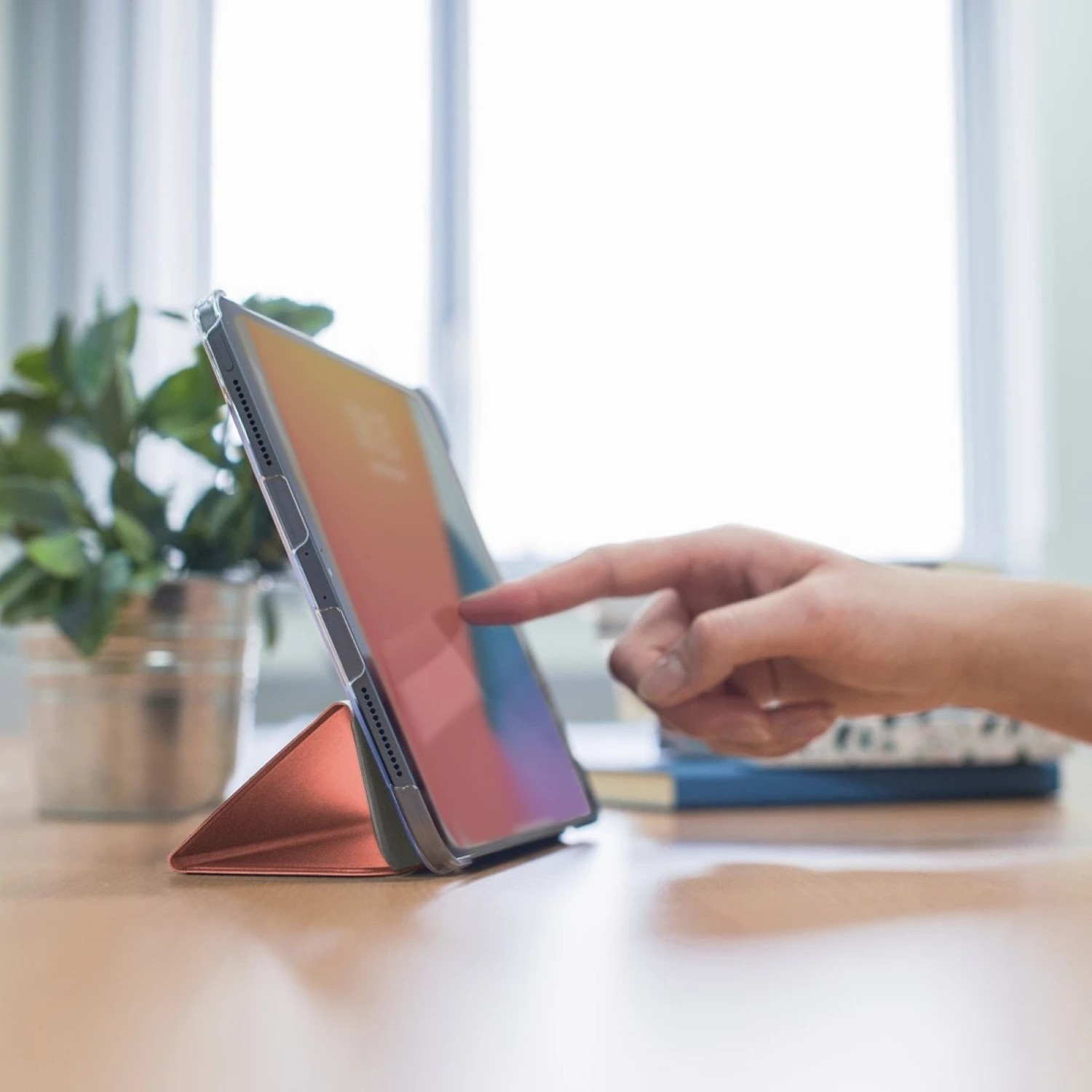 Ręka dotyka tabletu na stojaku. Roślina w srebrnej doniczce jest w tle. Widoczne są również książki.