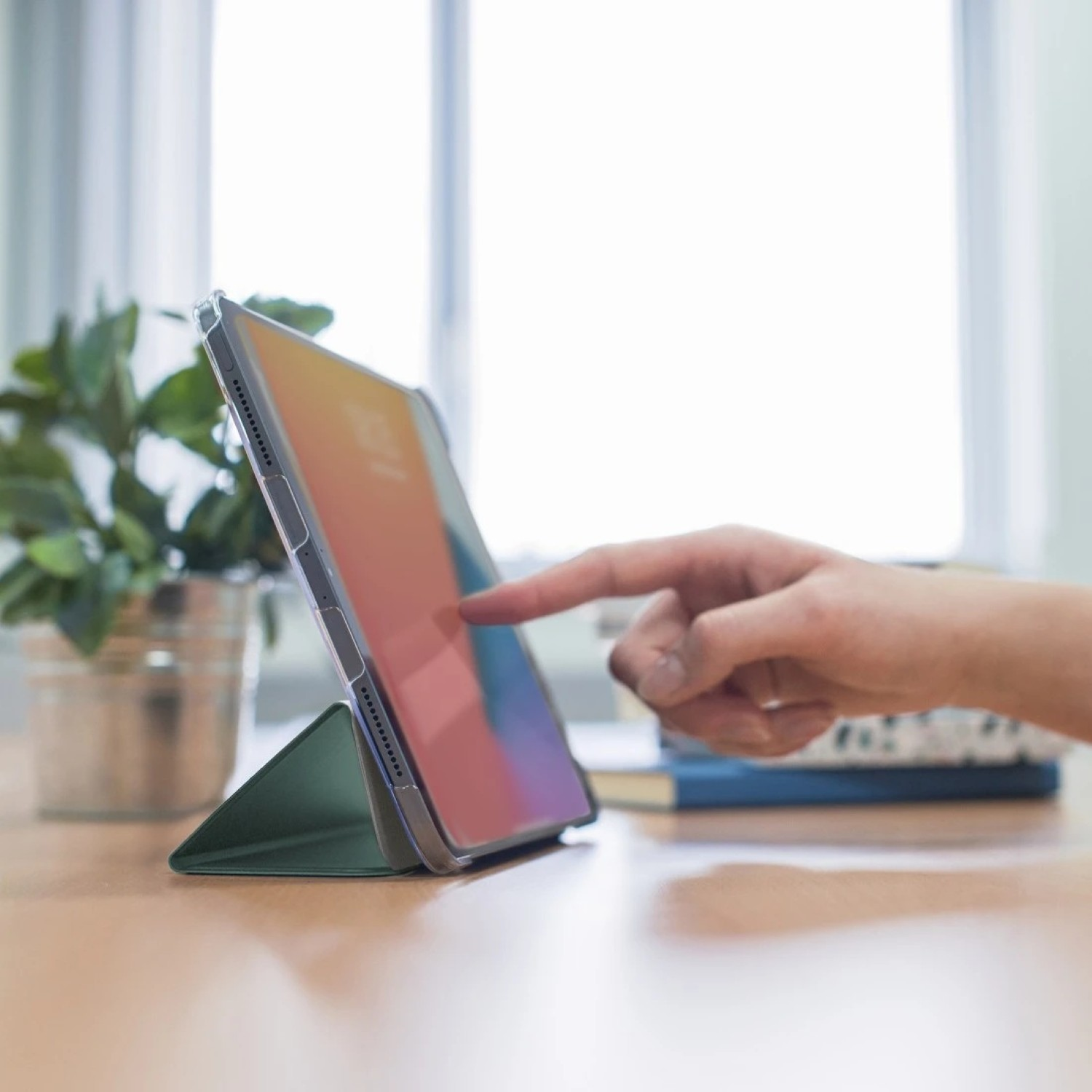 Ręka dotyka ekranu tabletu z kolorowym gradientem. Tablet jest podparty na zielonym stojaku. Roślina i książki w tle.