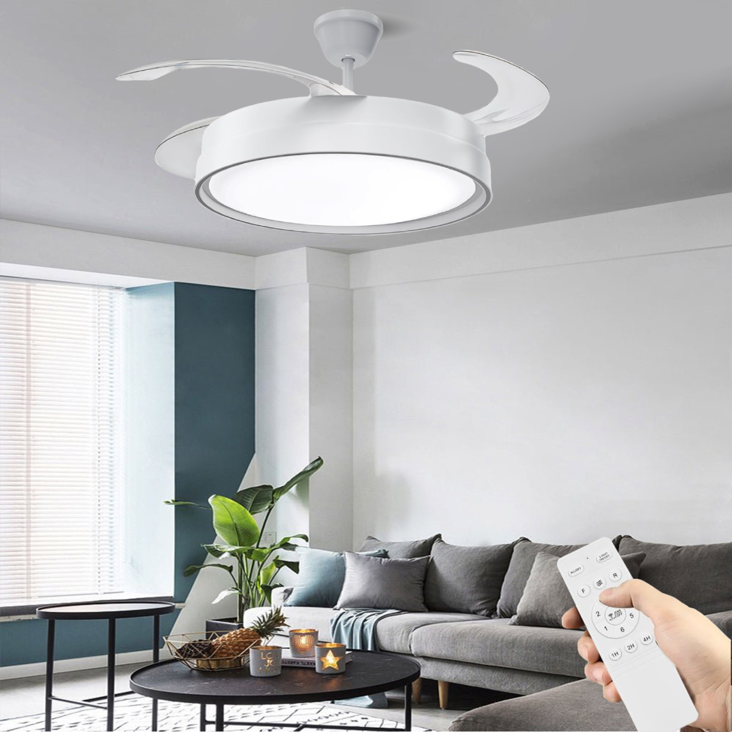 Un ventilador de techo blanco con luces está encendido. Una persona sostiene el control remoto. La habitación tiene un sofá, mesas y plantas.