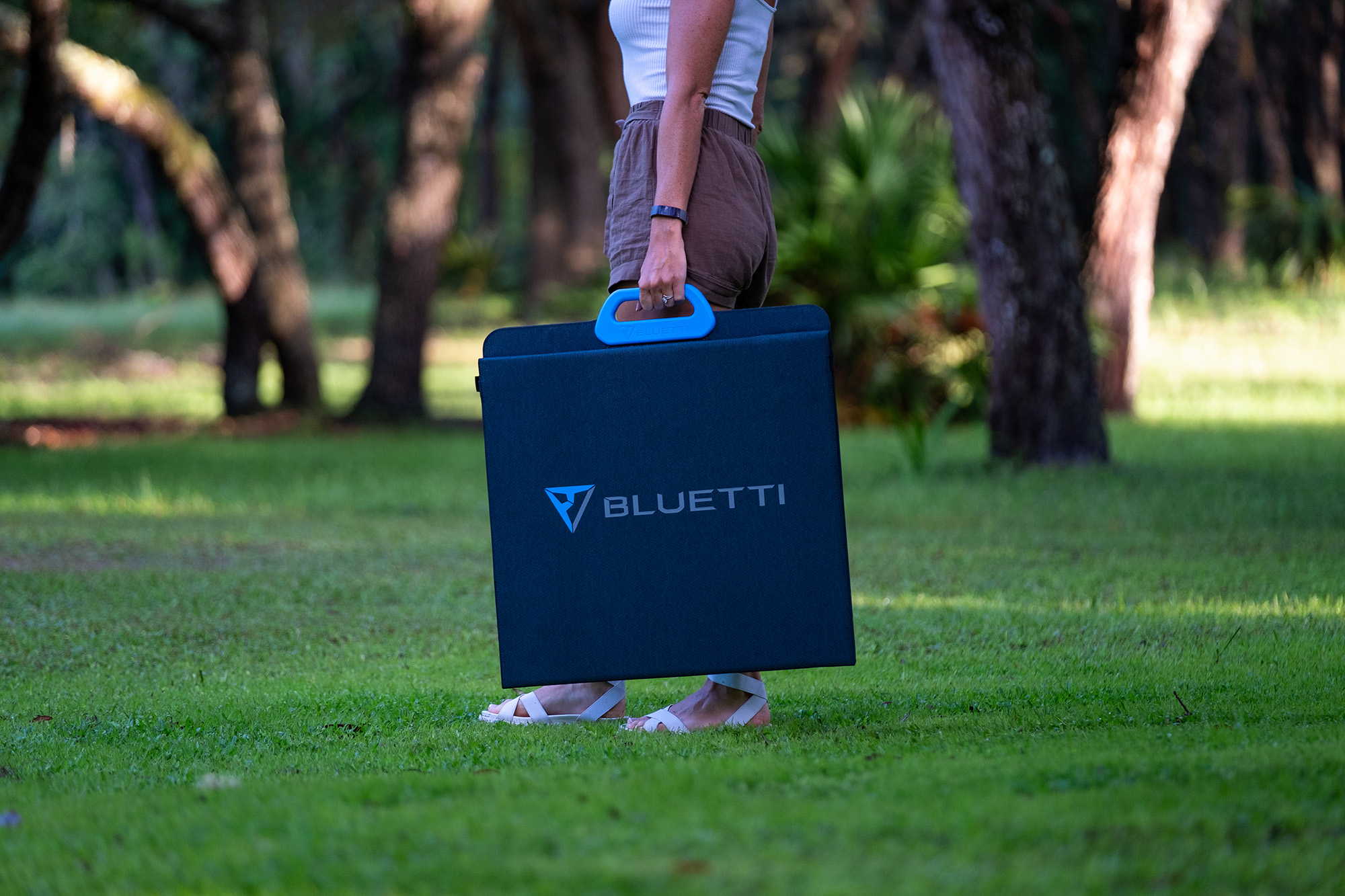 Persona sosteniendo una funda azul de Bluetti con asa azul, de pie sobre hierba verde. Árboles en el fondo.
