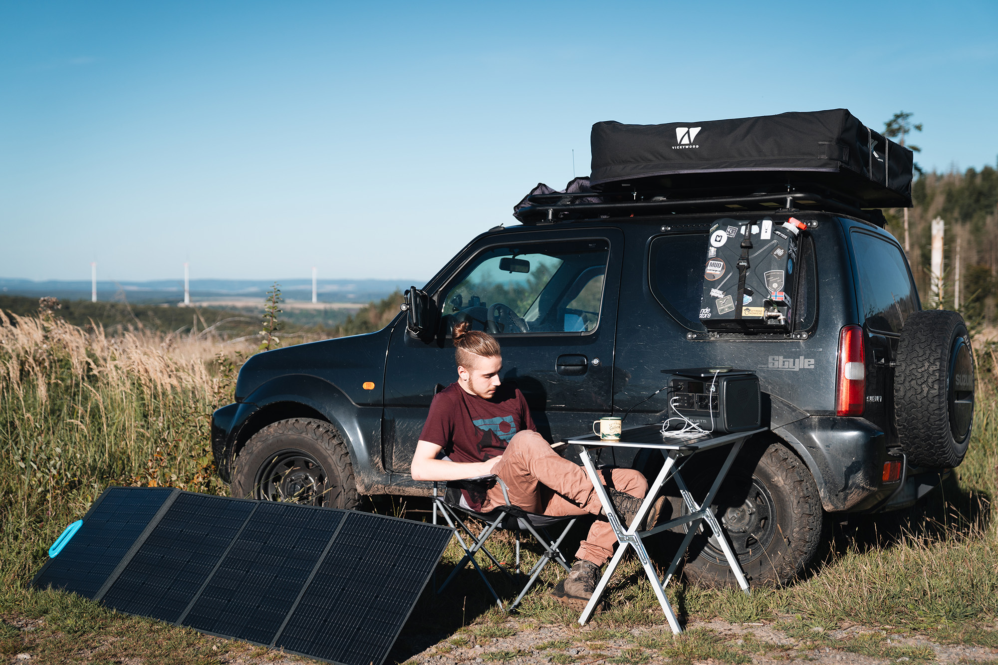 Un hombre sentado junto a un SUV negro en un campo. Paneles solares cerca, y el coche tiene una tienda de techo.