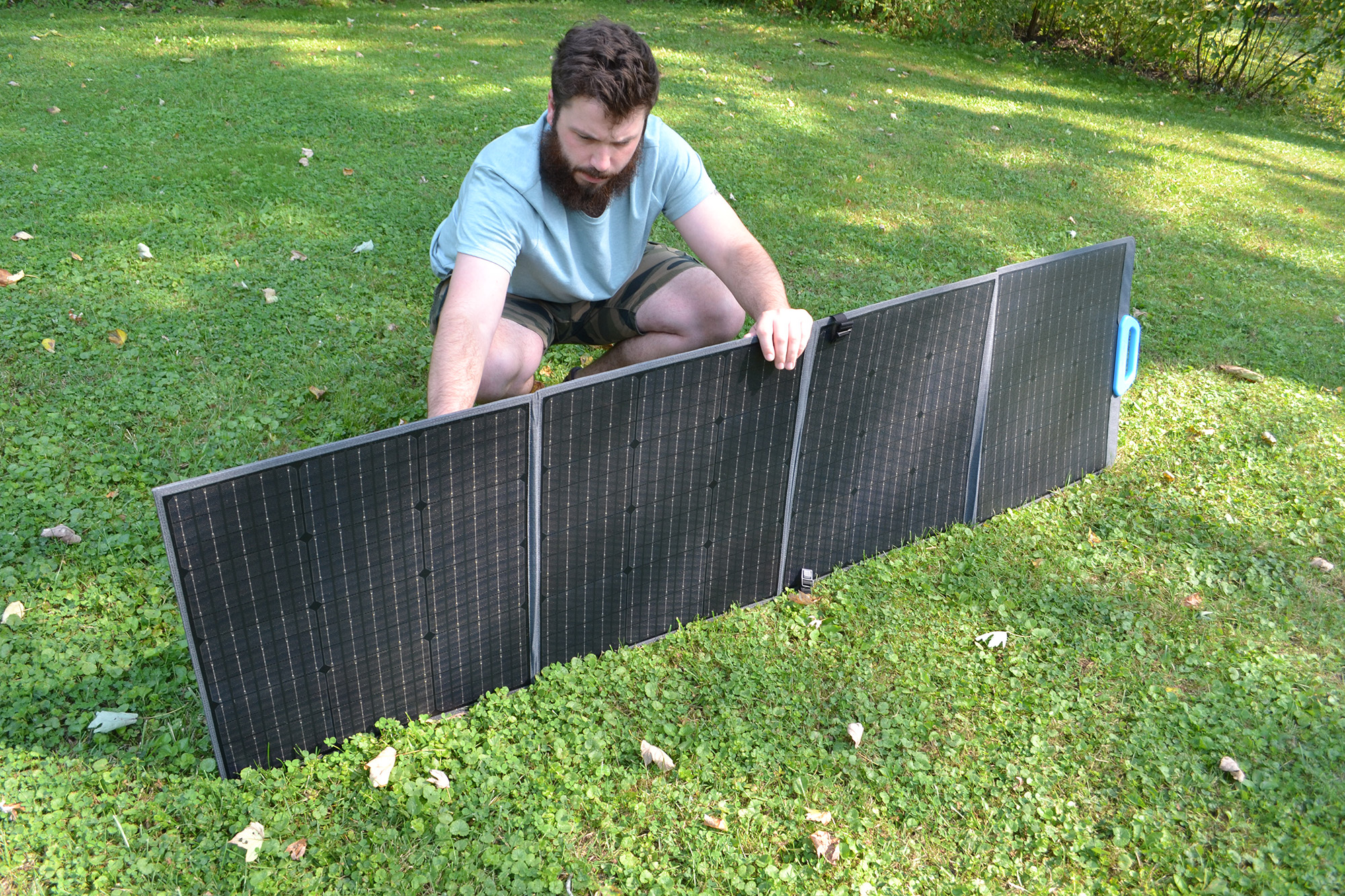 Un hombre se arrodilla en la hierba, instalando un panel solar plegable. El panel es negro con un asa azul. El fondo incluye árboles y hierba.