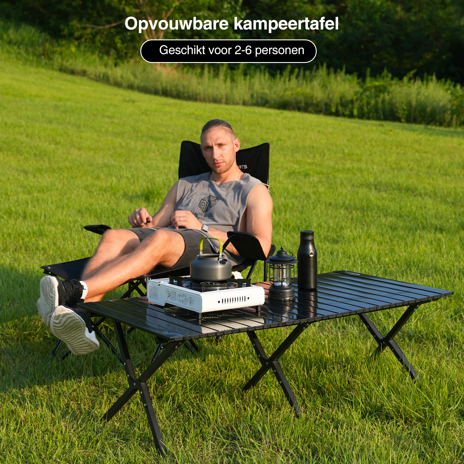 Man ontspant zich bij een campingtafel. Een waterkoker, een lantaarn en een waterfles staan op de tafel. Groen gras op de achtergrond.