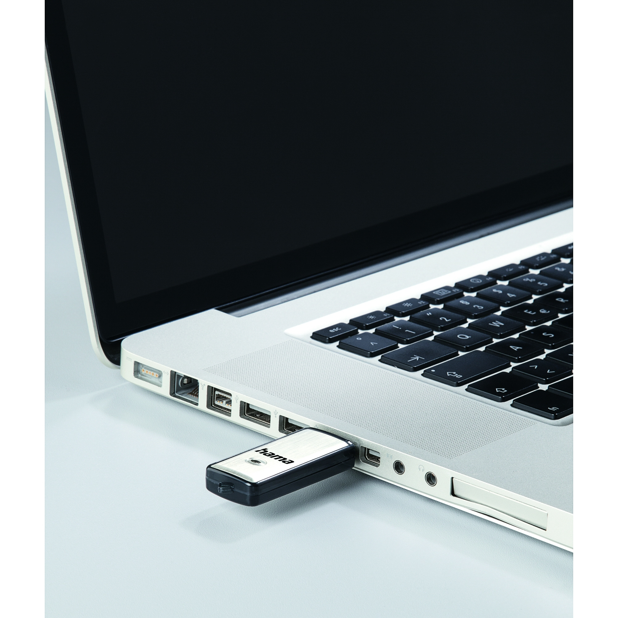 Ein silberner Laptop mit schwarzer Tastatur. Ein schwarzer und silberner USB-Stick steckt seitlich, mit der Aufschrift 'hama'.
