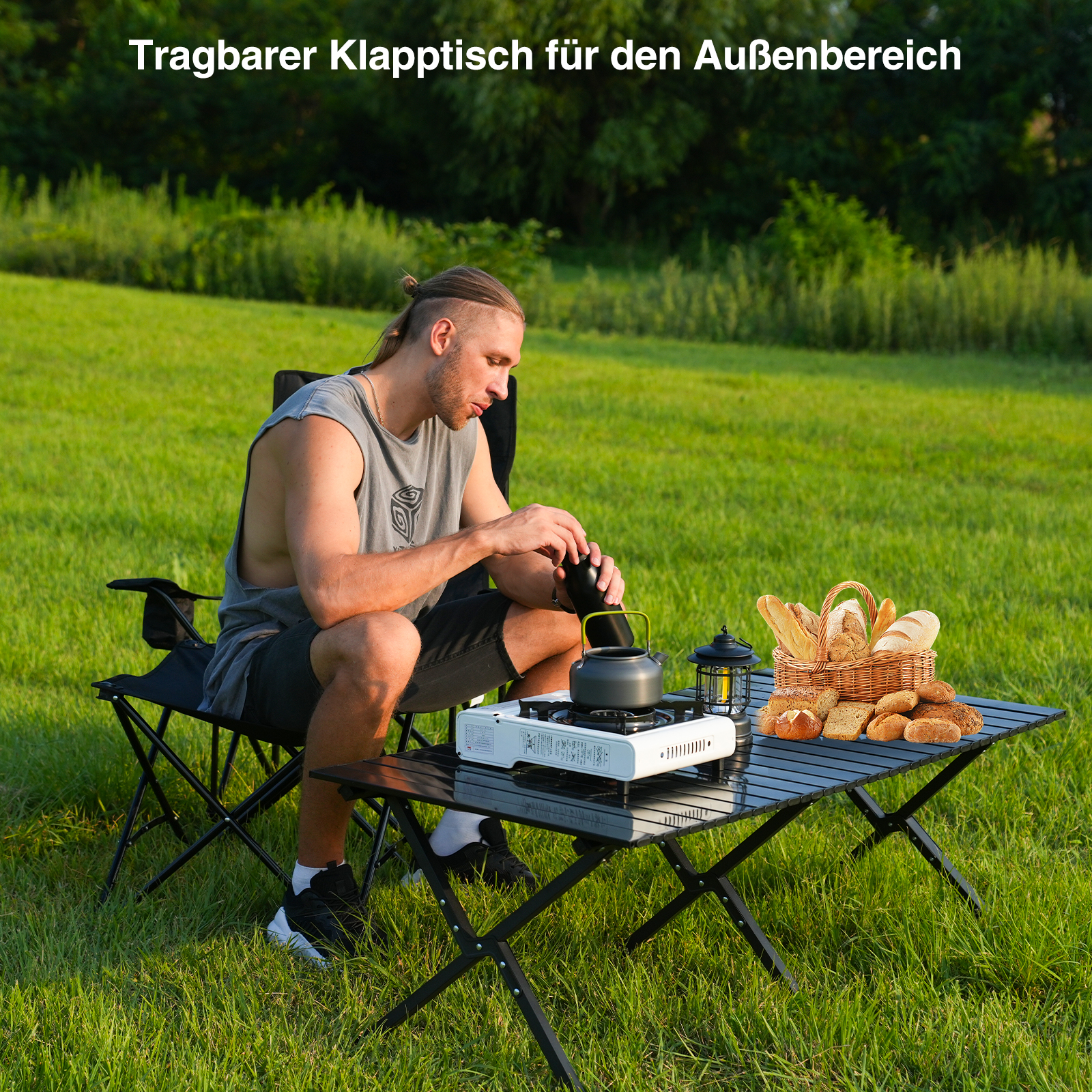 Ein Mann sitzt an einem Klapptisch mit einem Wasserkocher. Ein Brotkorb und eine Laterne stehen auf dem Tisch. Grünes Gras im Hintergrund.