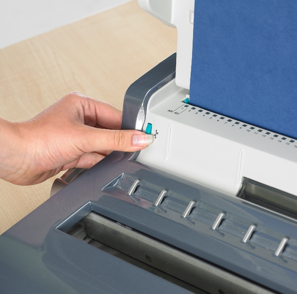 Eine Hand bedient eine Bindemaschine. Ein blauer Papierstapel befindet sich in der Maschine. Graue und weiße Maschinenteile sind sichtbar.