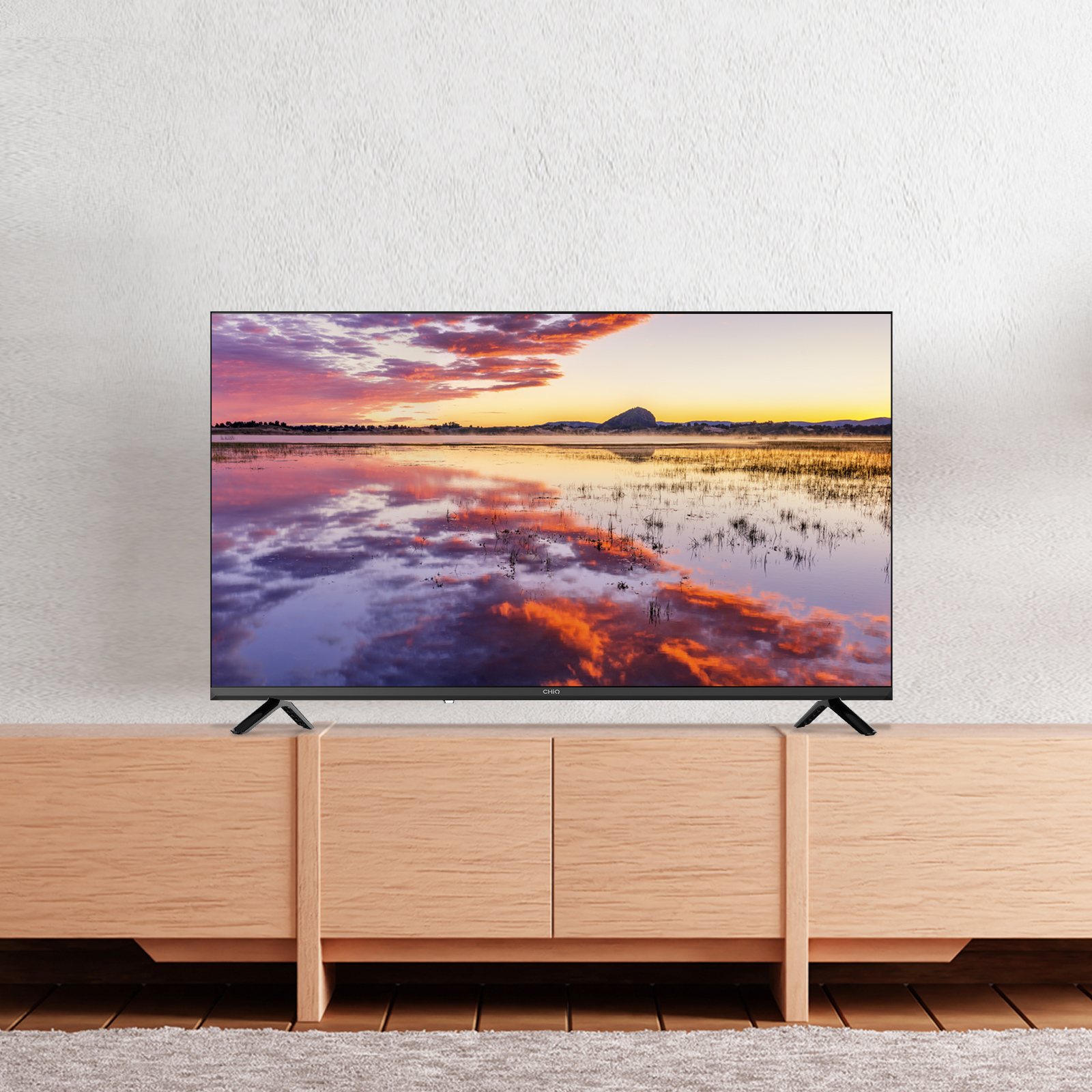 Telewizor wyświetla zachód słońca nad wodą. Stoi na drewnianej szafce. W tle biała, teksturowana ściana.