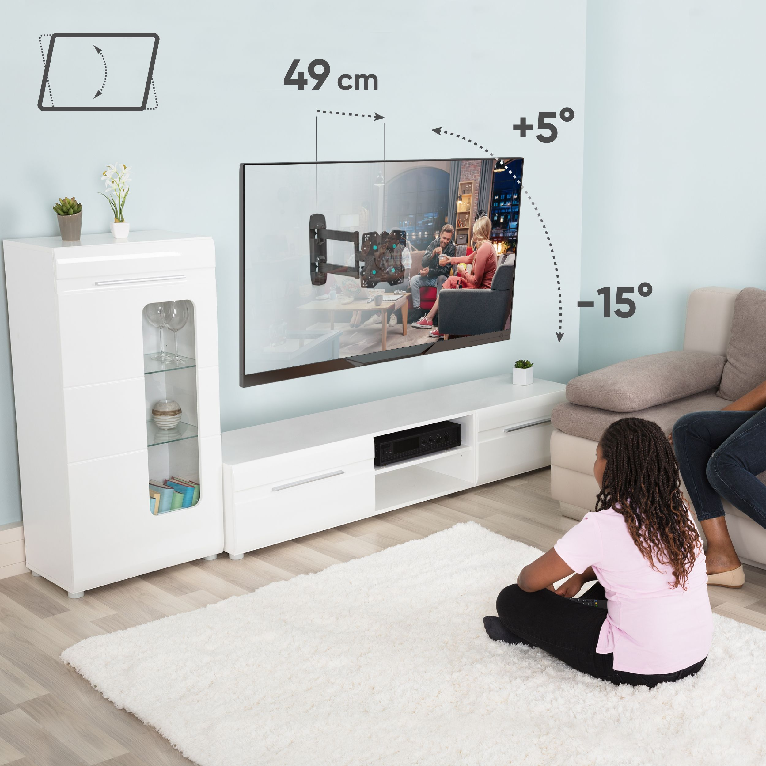 Telewizor jest zamontowany na jasnoniebieskiej ścianie. Kobieta siedzi na białym dywanie i ogląda telewizję. Szafka jest po lewej stronie.