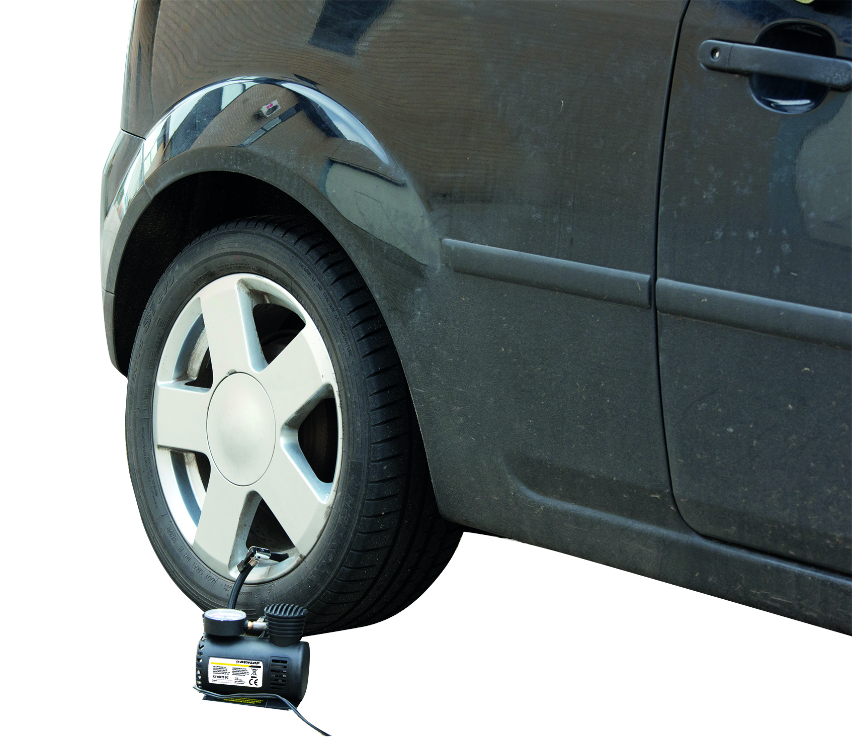 Pneumatico nero di un'auto con cerchio argentato. Un compressore d'aria nero è collegato al pneumatico, gonfiandolo. L'auto è sporca.