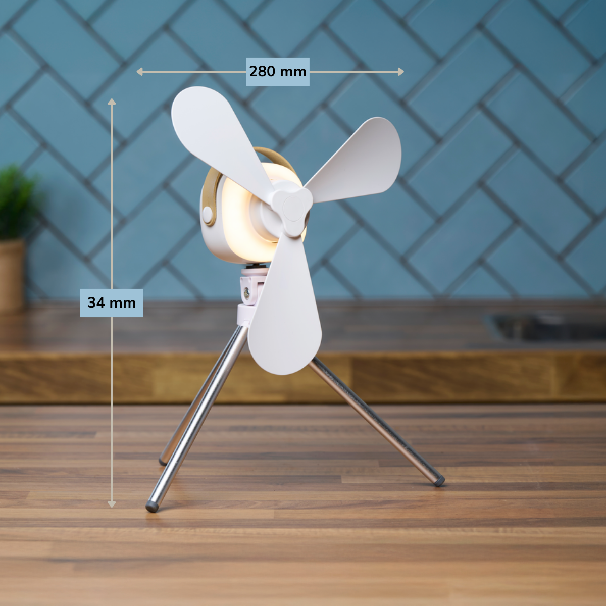 Un mini ventilateur blanc et rose sur un trépied, avec une lumière brillante. Il est sur une surface en bois, avec un fond de carreaux bleus.