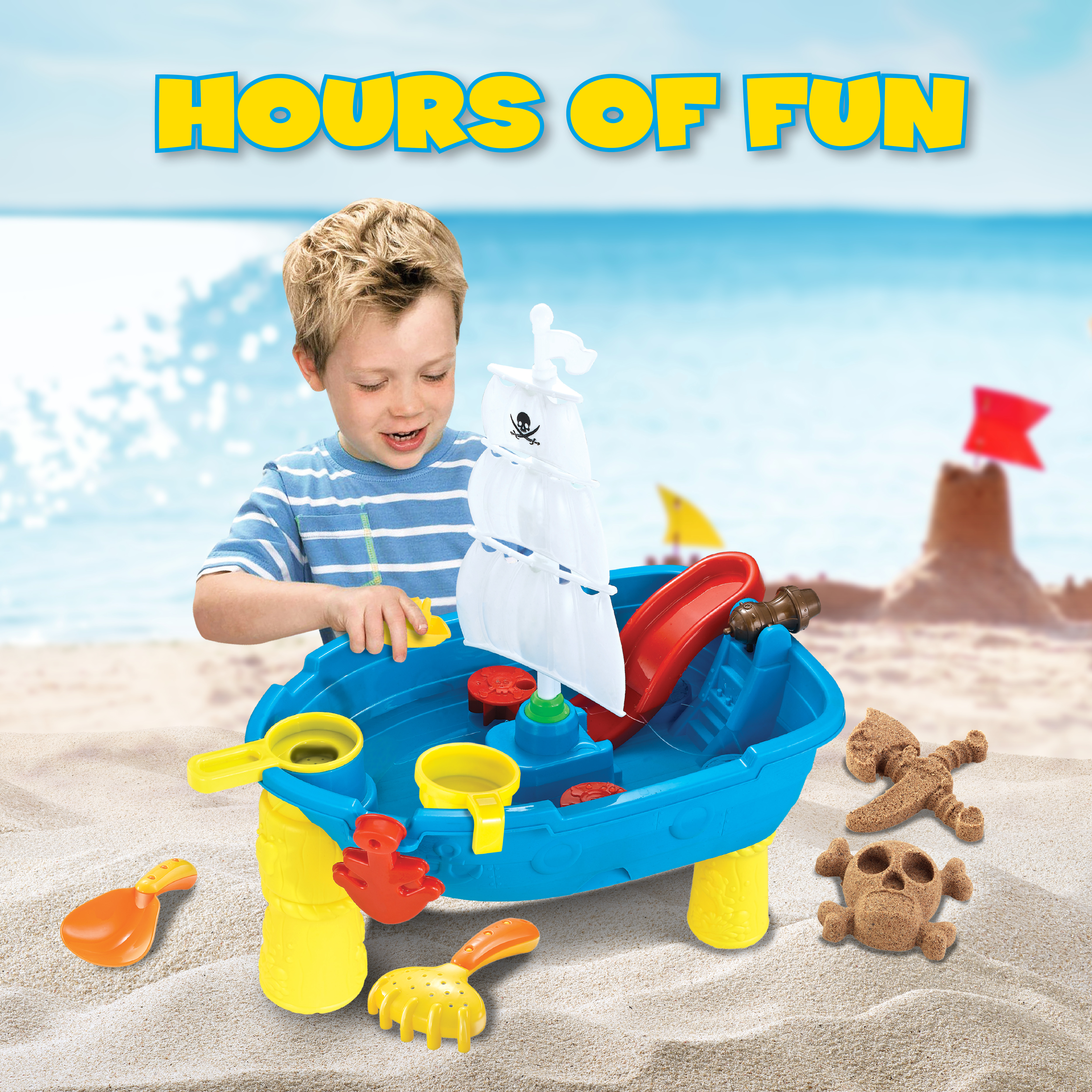 A boy plays with a pirate ship sand table on a beach. Sand tools and sand sculptures are nearby. The sky is blue.