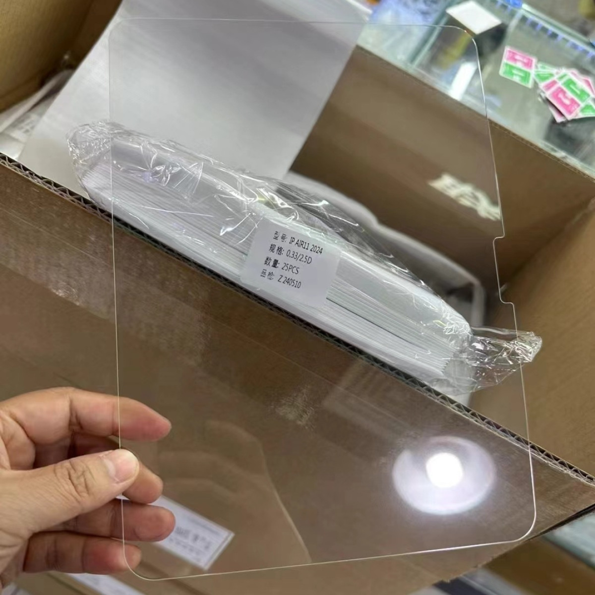 Hand houdt een transparante schermbeschermer vast, naast een stapel beschermers in plastic verpakt in een kartonnen doos.