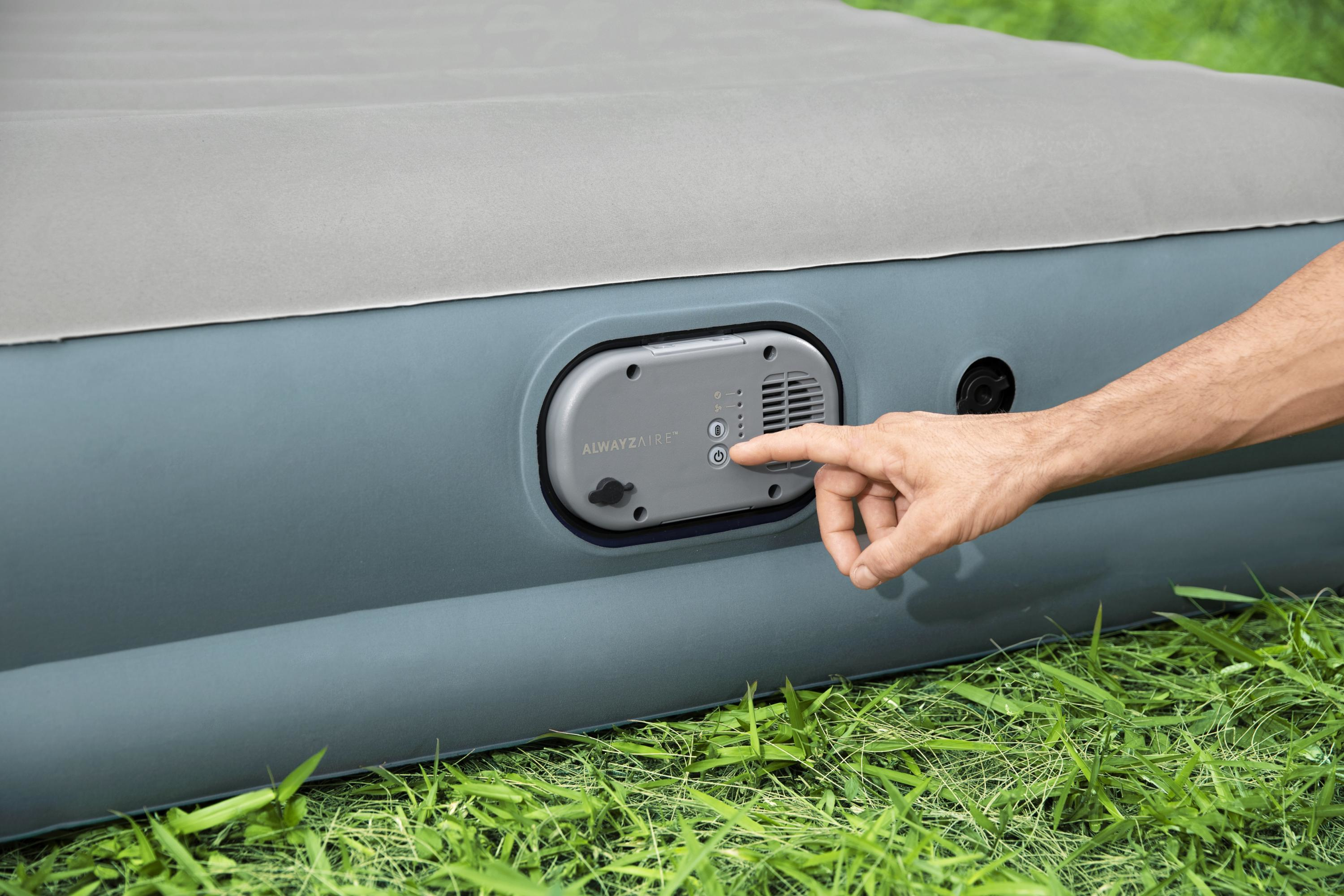 Een hand drukt op een knop op het bedieningspaneel van een luchtmatras. De matras ligt op gras.