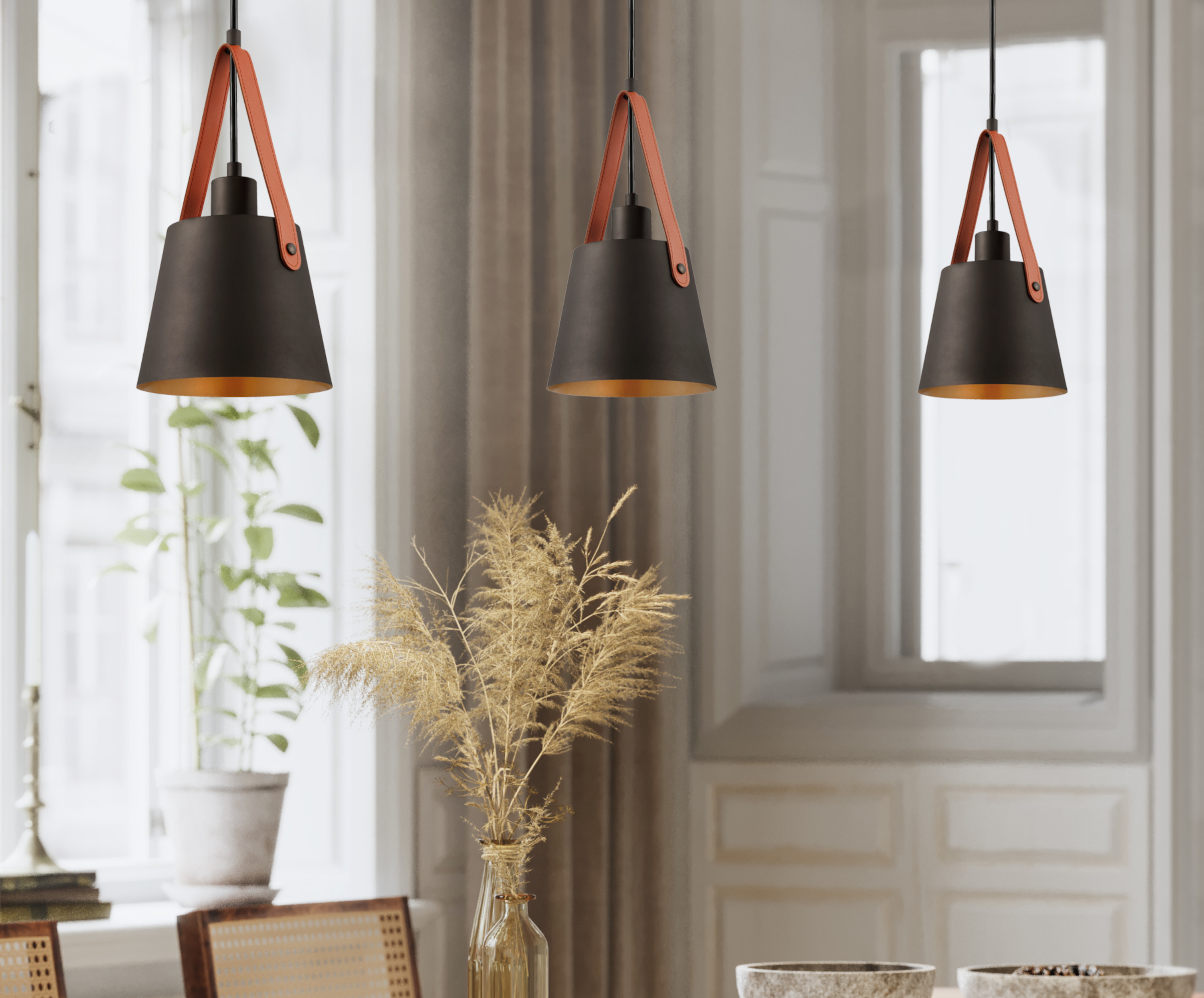 Drei hängende schwarze Lampen mit orangefarbenen Lederriemen. Eine Vase mit getrockneten Blumen und eine Pflanze im Topf stehen vor einem Fenster.