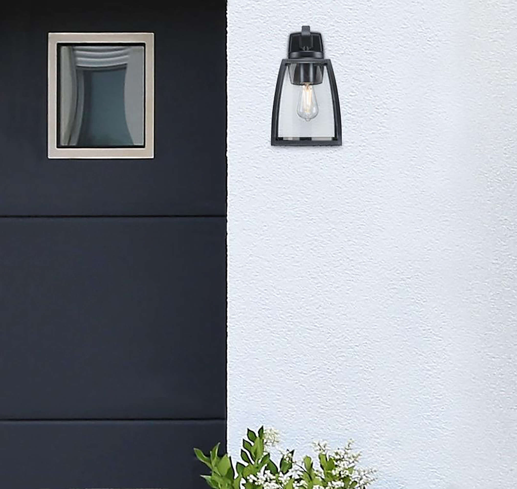 Schwarze Außenlampe an weißer Wand. Tür mit Fenster links. Grüne Blätter und weiße Blumen unten.