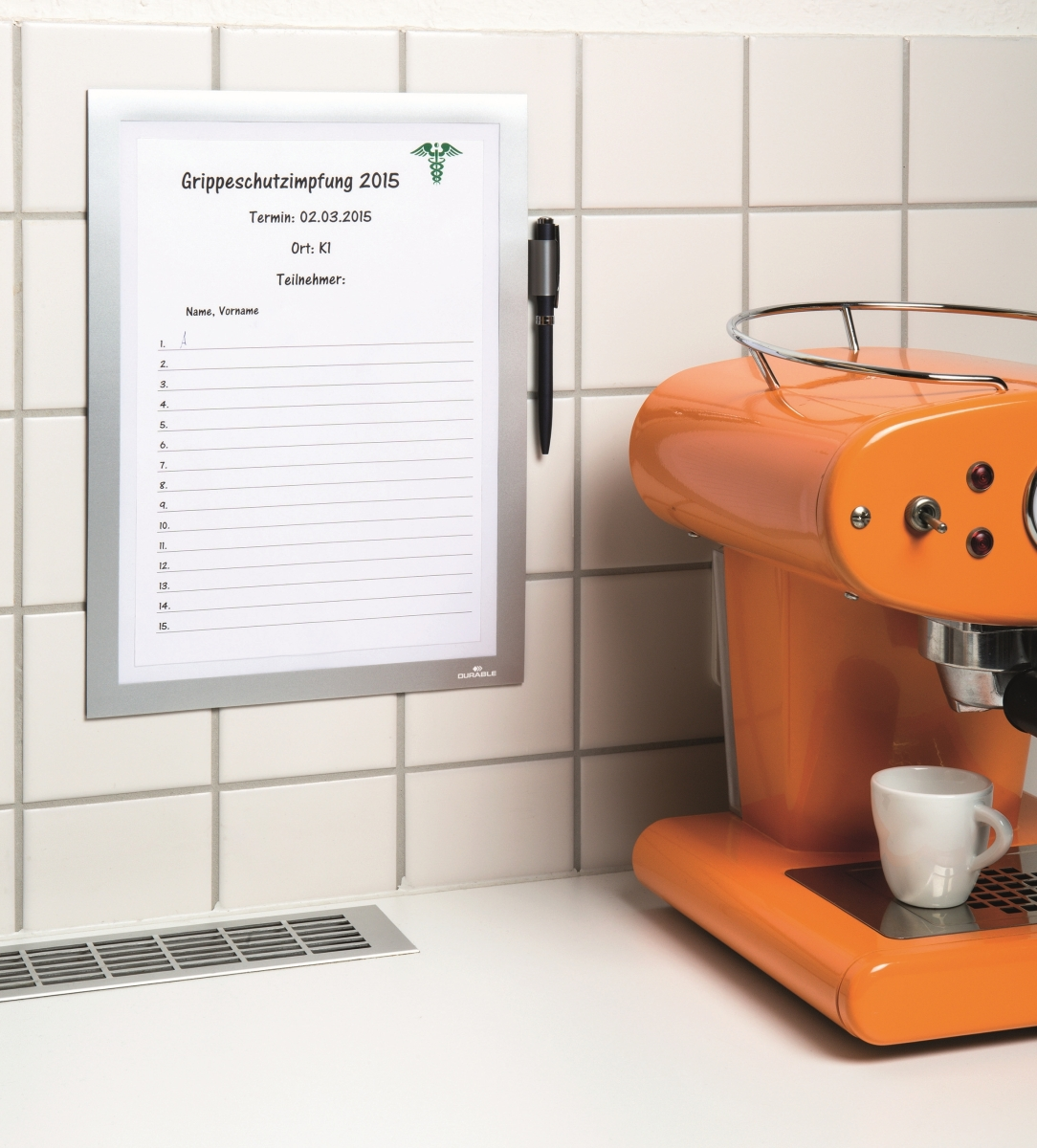 Eine weiße Tafel listet Namen für eine Grippeschutzimpfung 2015 auf, daneben hängt ein schwarzer Stift, daneben steht eine orangefarbene Espressomaschine.