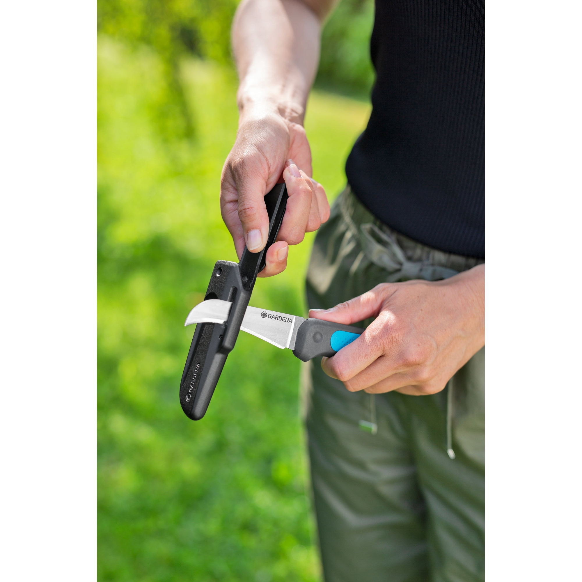 Una persona tiene in mano un coltello con un fodero nero, lo sta rimuovendo. Il coltello ha un manico blu e grigio. Sfondo verde.