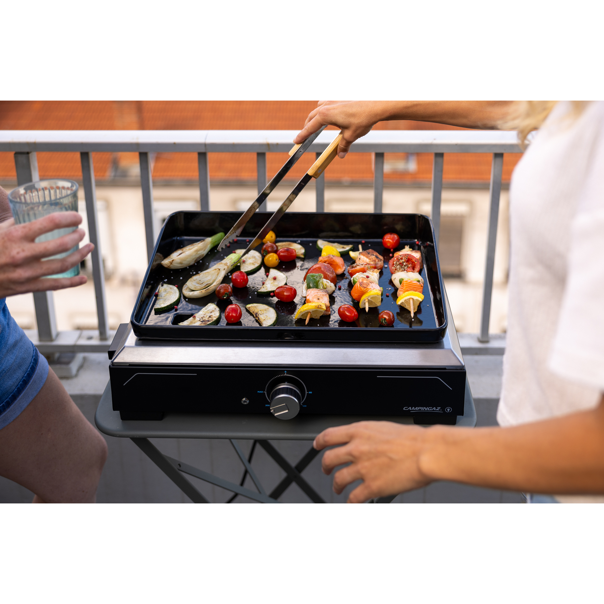 Eine Person grillt Gemüse und Spieße auf einem schwarzen Grill. Eine andere Person hält ein Glas Wasser.