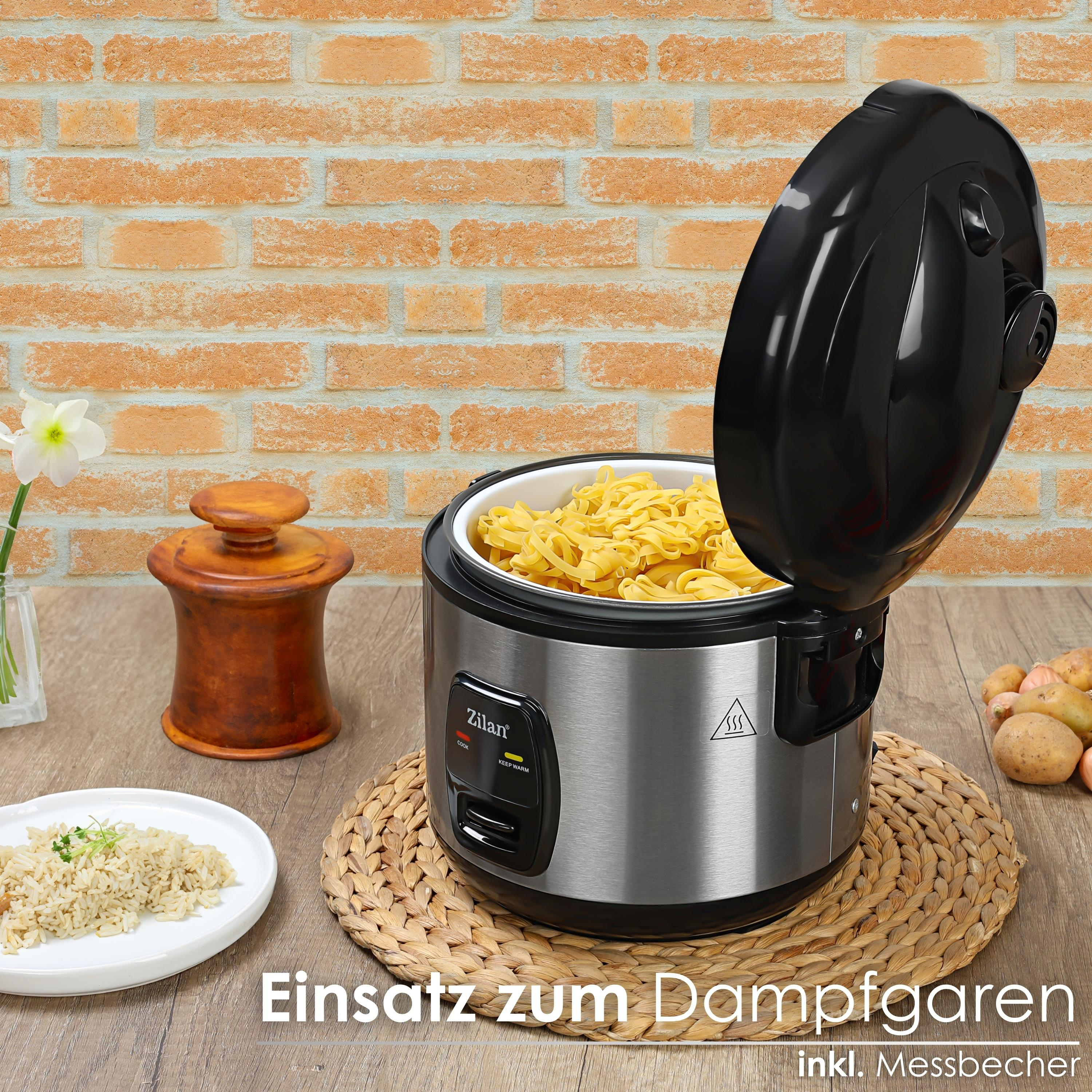 Reiskocher mit Nudeln. Ein Holztisch, ein Teller mit Reis und ein Holzgefäß stehen auf dem Tisch. Eine Backsteinmauer im Hintergrund.