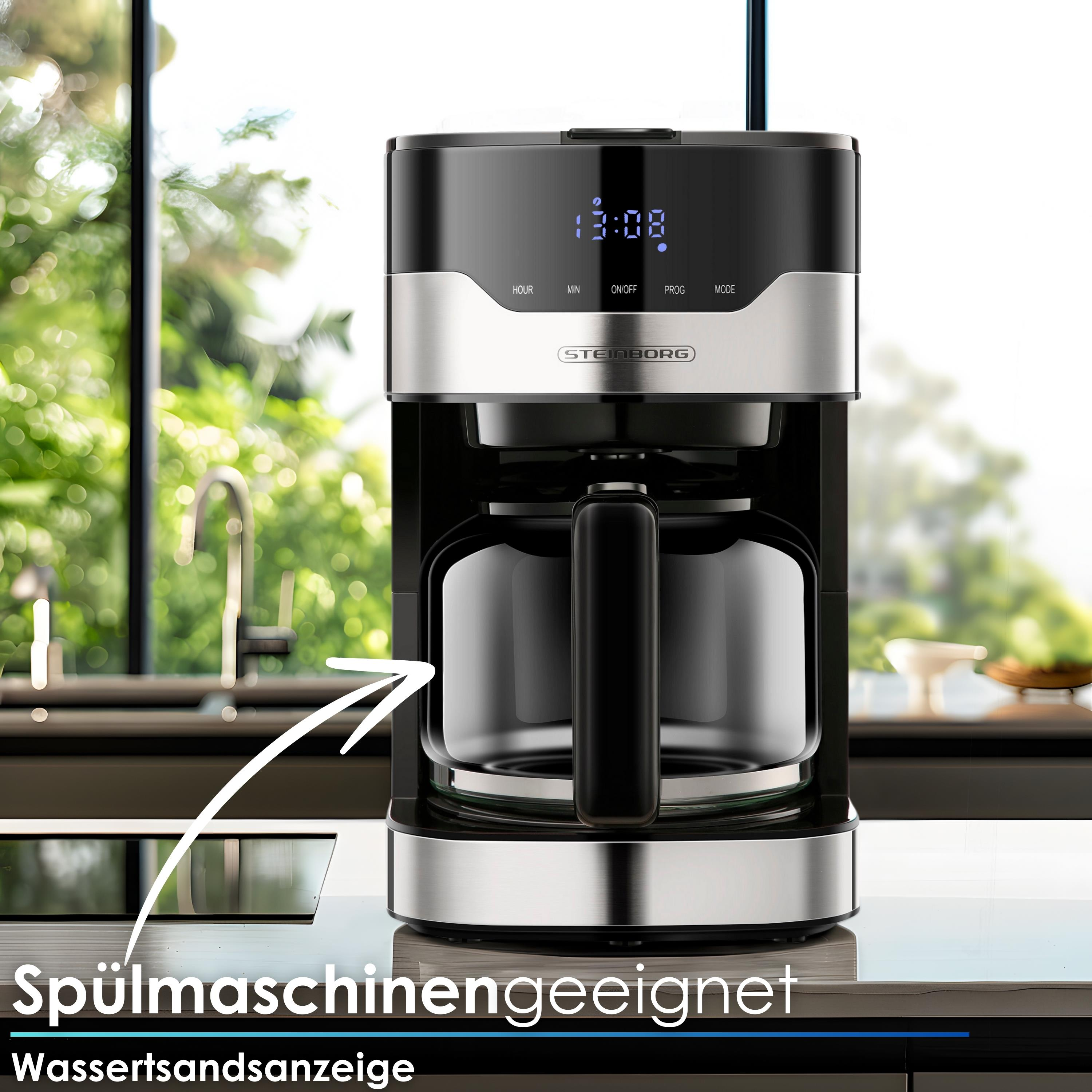 Eine schwarz-silberne Kaffeemaschine mit Glaskaraffe steht auf einer Arbeitsplatte. Grünes Laub ist durch ein Fenster sichtbar.